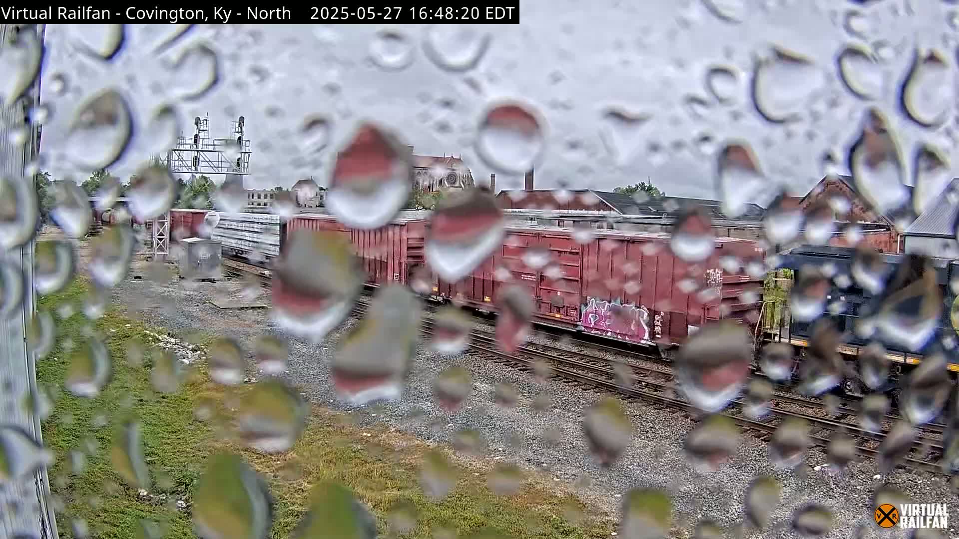 A view of railroad cars and tracks seen through a rain-covered window during a rainy day.