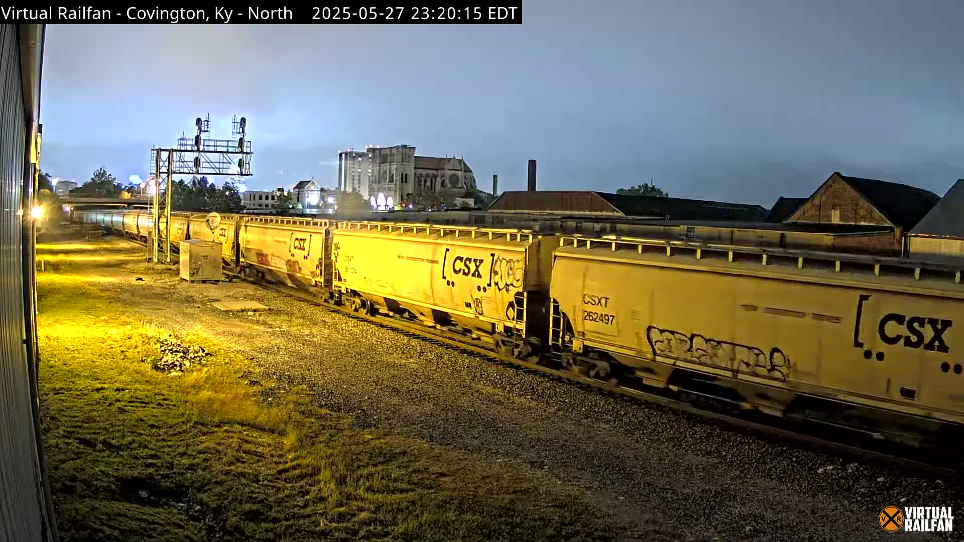 A long freight train of yellow CSX cars moves along the tracks at night under a twilight sky.