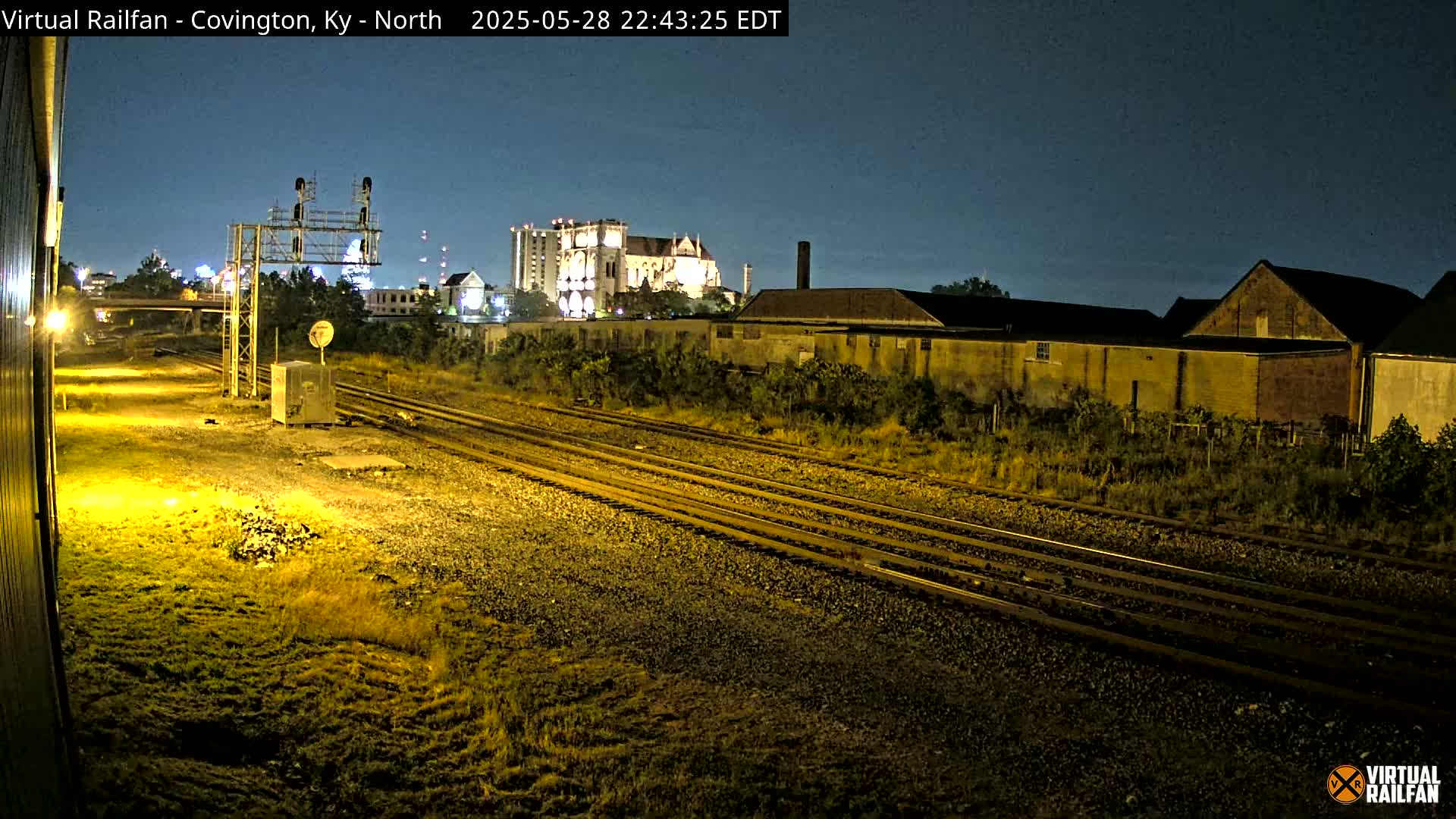 A nighttime view of multiple train tracks running alongside industrial buildings and a taller building in the background under a clear, dark sky.