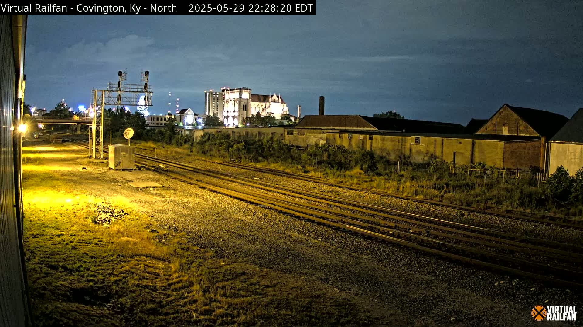 Under a dark, mostly clear night sky, several sets of train tracks run through a weedy, gravel area past industrial buildings and a large, illuminated building in the distance.