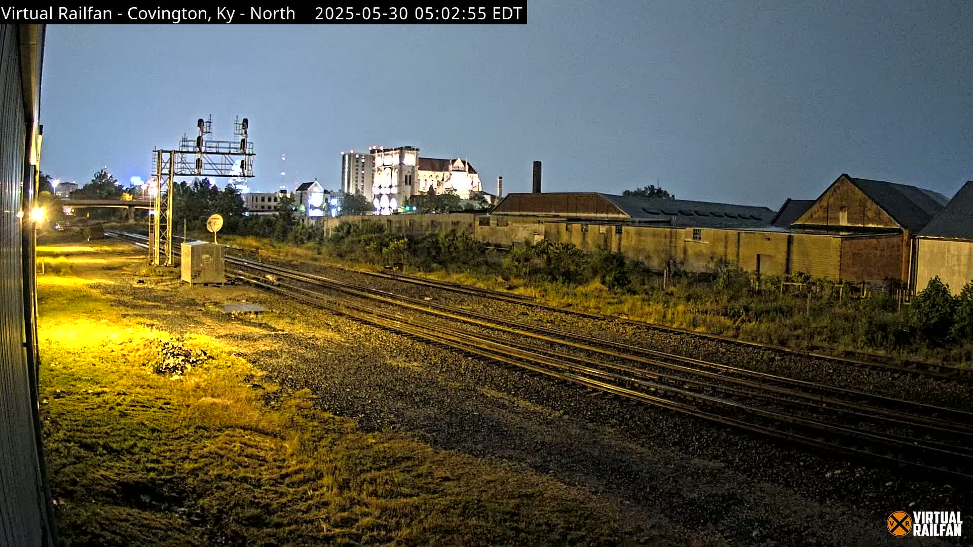 Under a dark, nighttime sky, railroad tracks stretch into the distance past industrial buildings and a taller structure that appears to be a church.