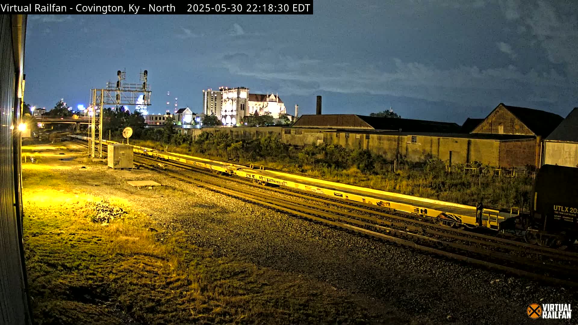 A nighttime view of a long freight train on multiple tracks, passing by industrial buildings under a partly cloudy sky.