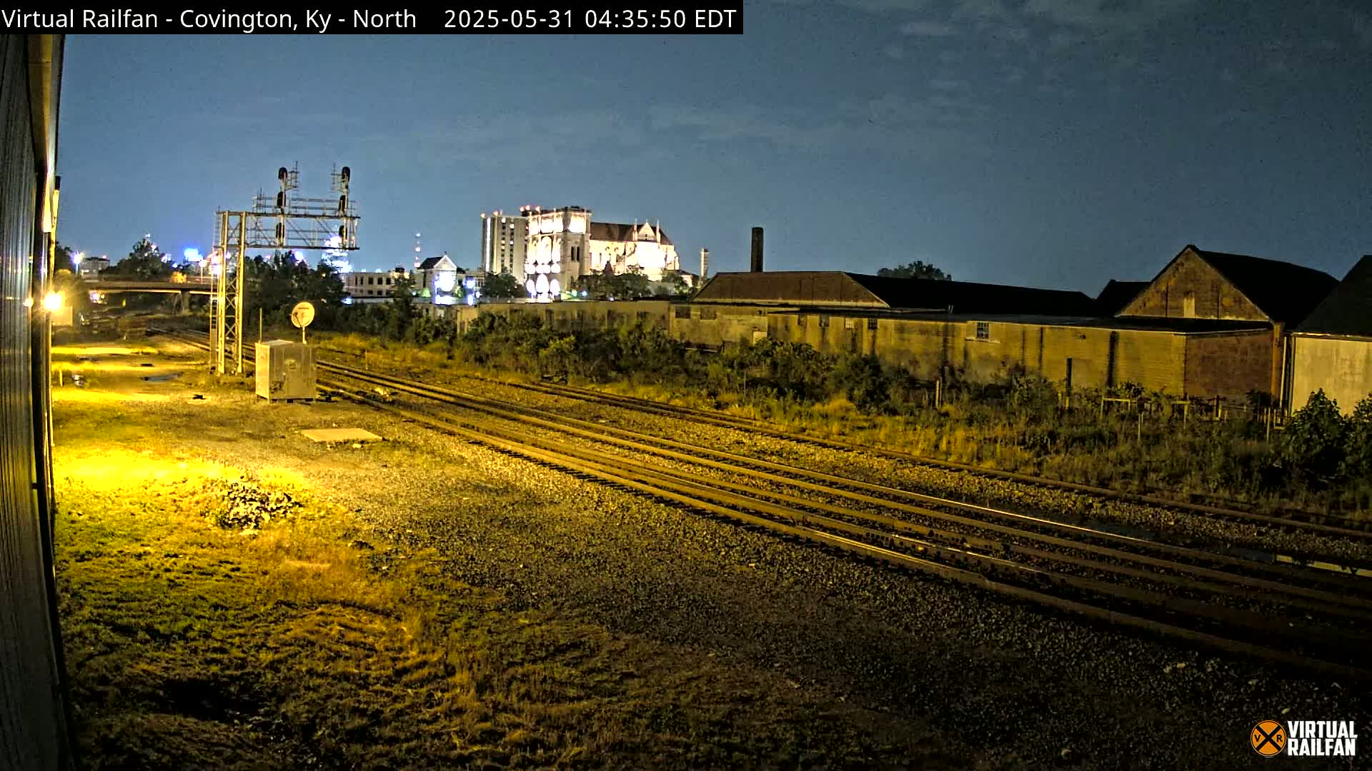 At night, under a mostly clear sky, multiple train tracks run through a weedy area past buildings and a large structure that may be a church.