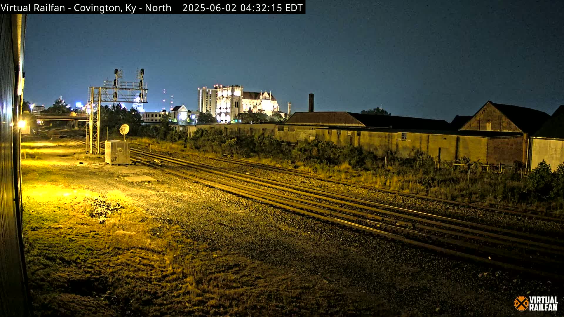 At night, under a clear sky, multiple sets of train tracks stretch toward a cityscape featuring a large building, smaller buildings, and vegetation.