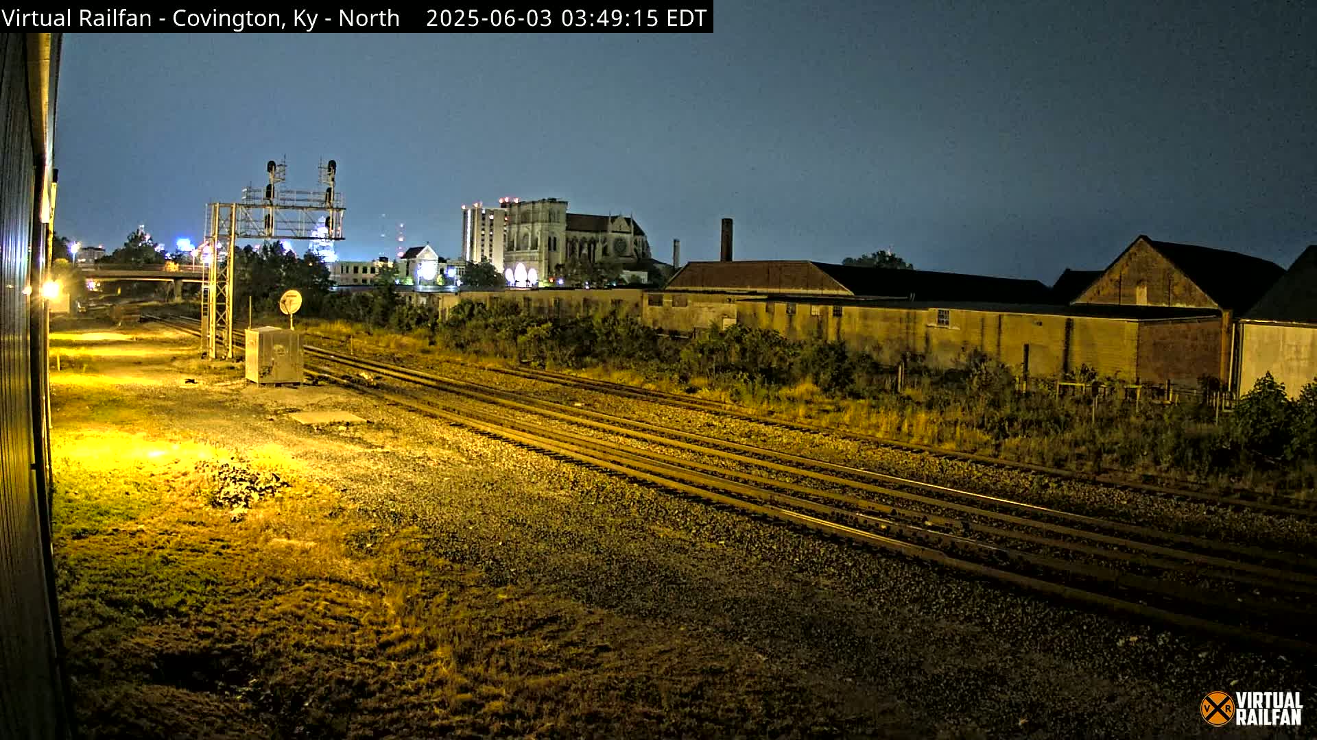 A nighttime view of multiple train tracks next to industrial buildings and a large cathedral under a dark sky.