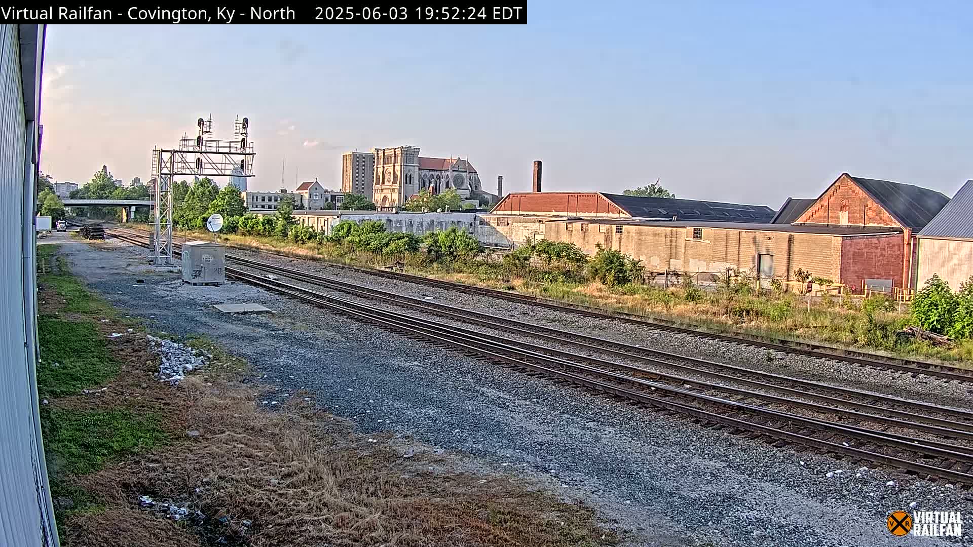 Under a clear sky, multiple sets of train tracks stretch toward a cityscape featuring industrial buildings and a large cathedral.