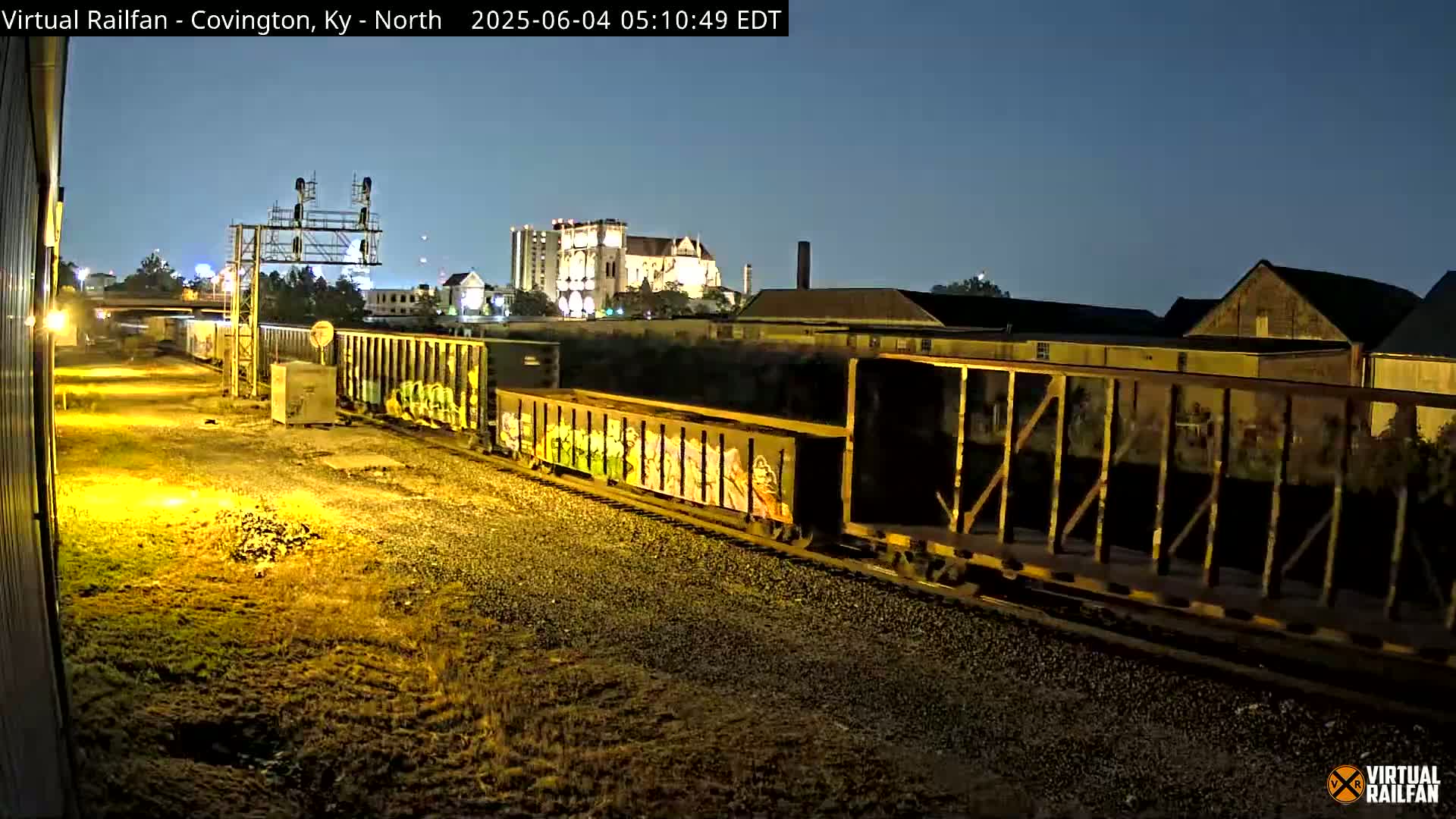 At night, a long freight train with graffiti-covered cars moves along railroad tracks past industrial buildings under a twilight sky.