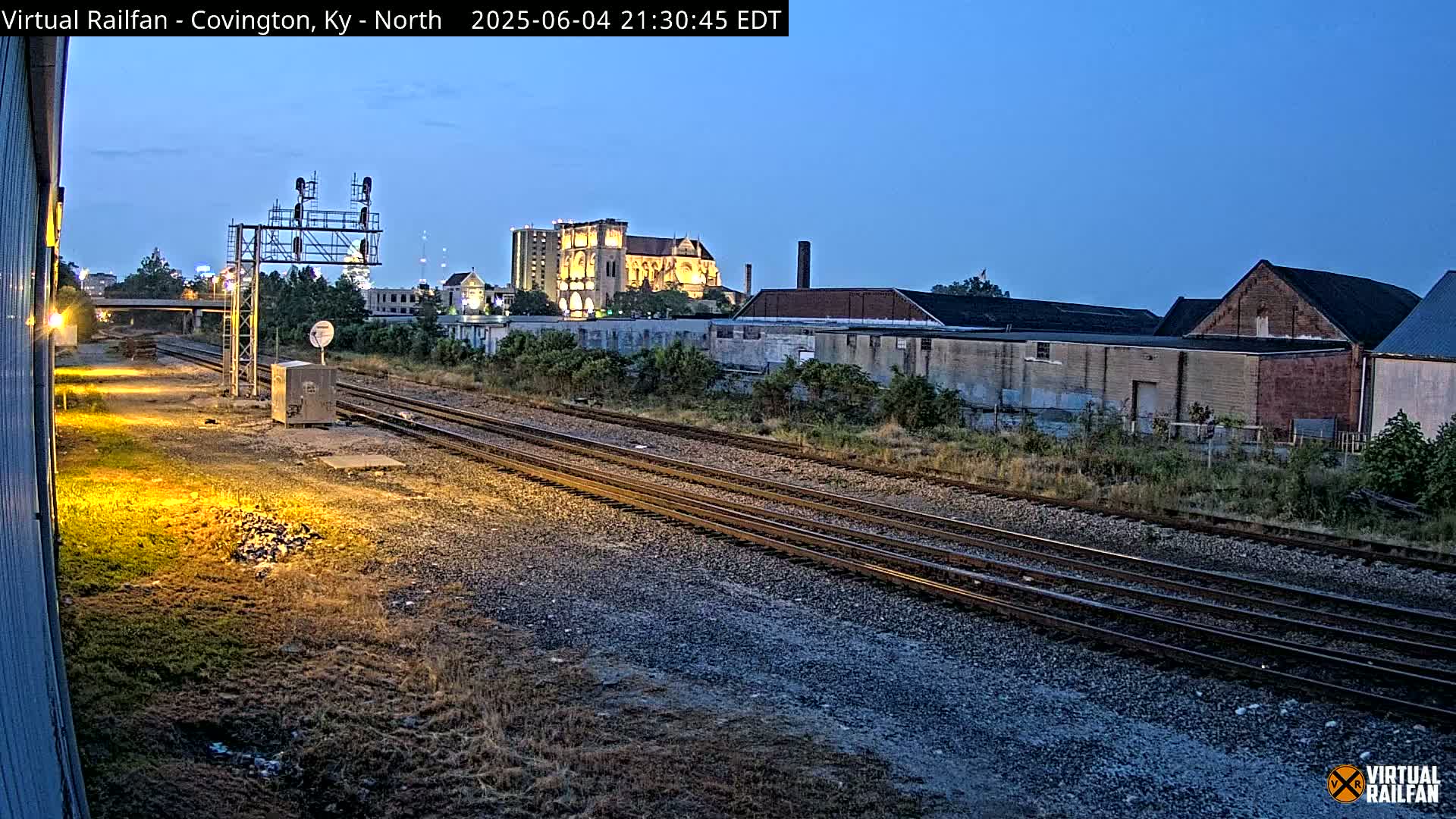 Train tracks at dusk, under a twilight sky, lead toward a city skyline featuring a large illuminated building.