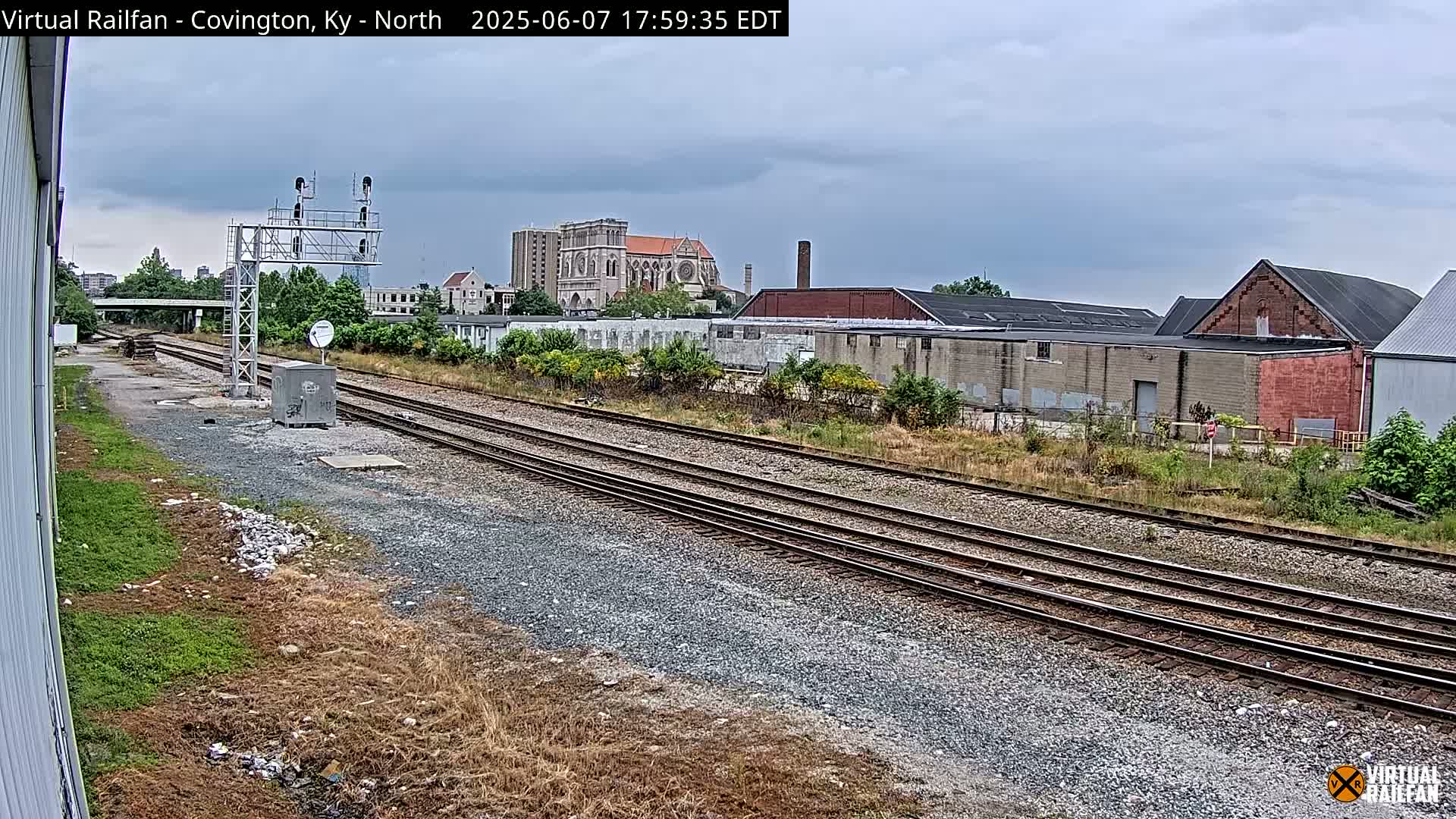 Multiple sets of train tracks extend into the distance under an overcast sky, past industrial buildings and a large church.