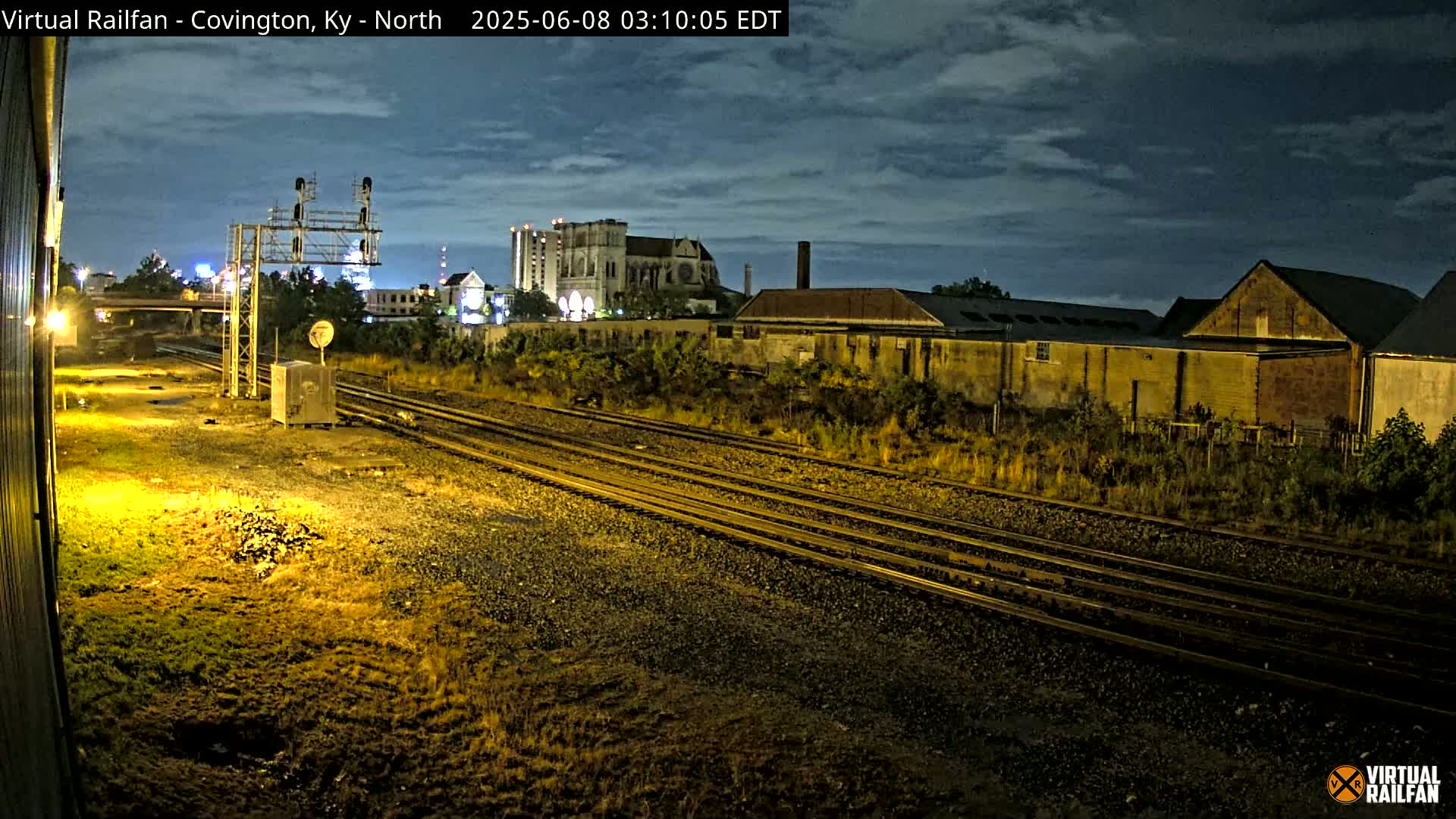 At night, under a partly cloudy sky, multiple train tracks run alongside buildings and vegetation.