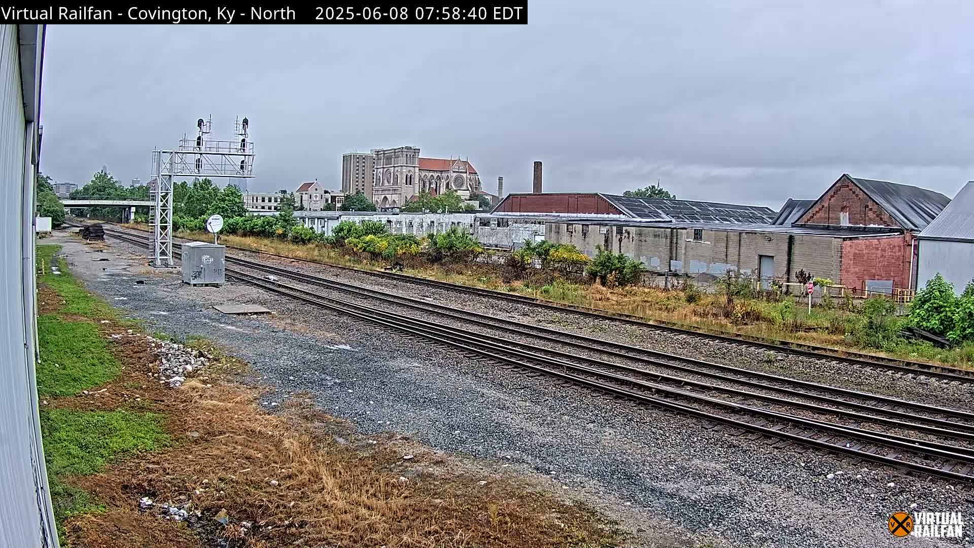 Multiple sets of train tracks run through an industrial area under an overcast sky, with a large cathedral visible in the background.