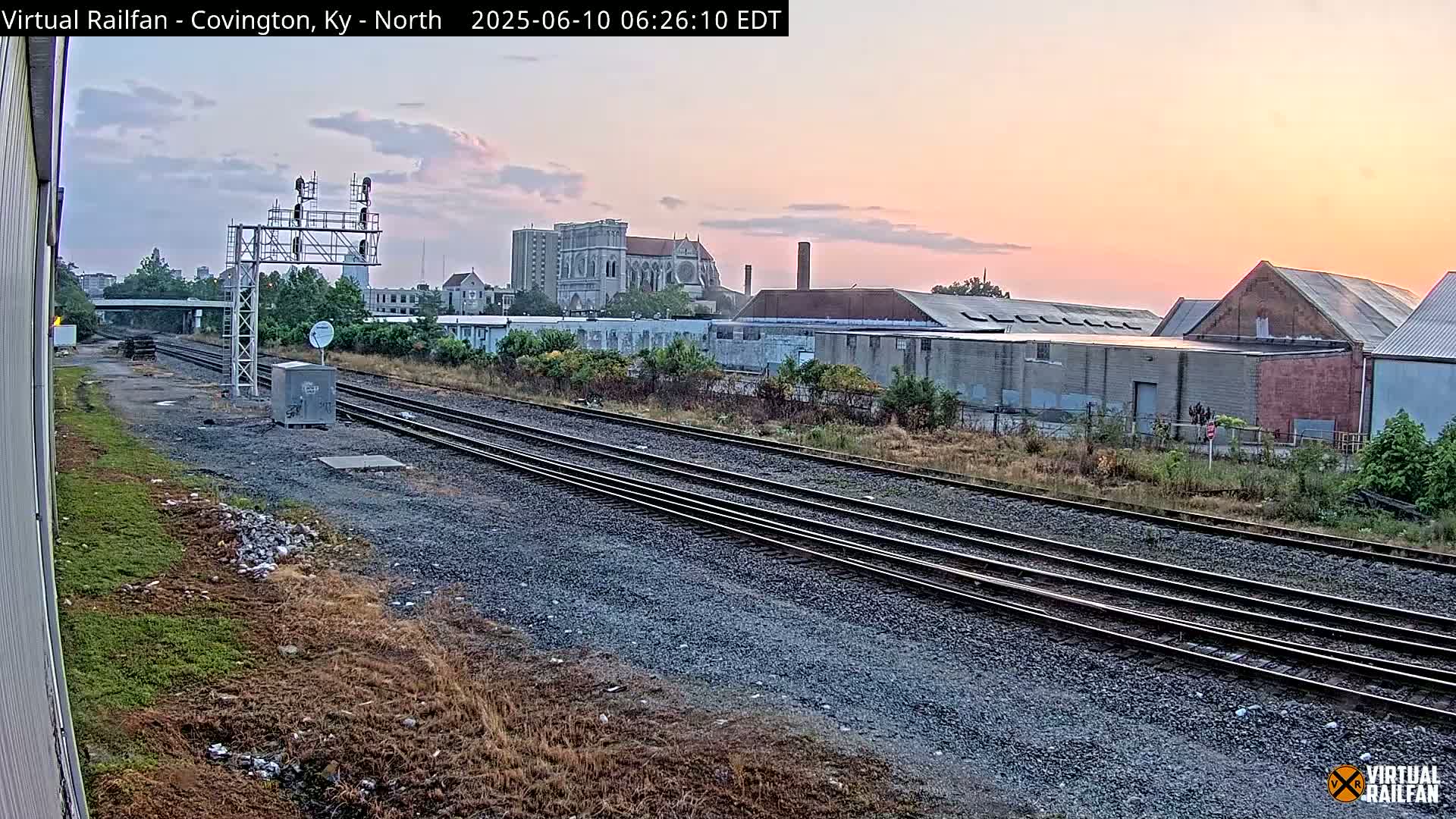 Multiple train tracks extend into the distance under a pale pink and orange sunrise, passing by industrial buildings and a large cathedral.