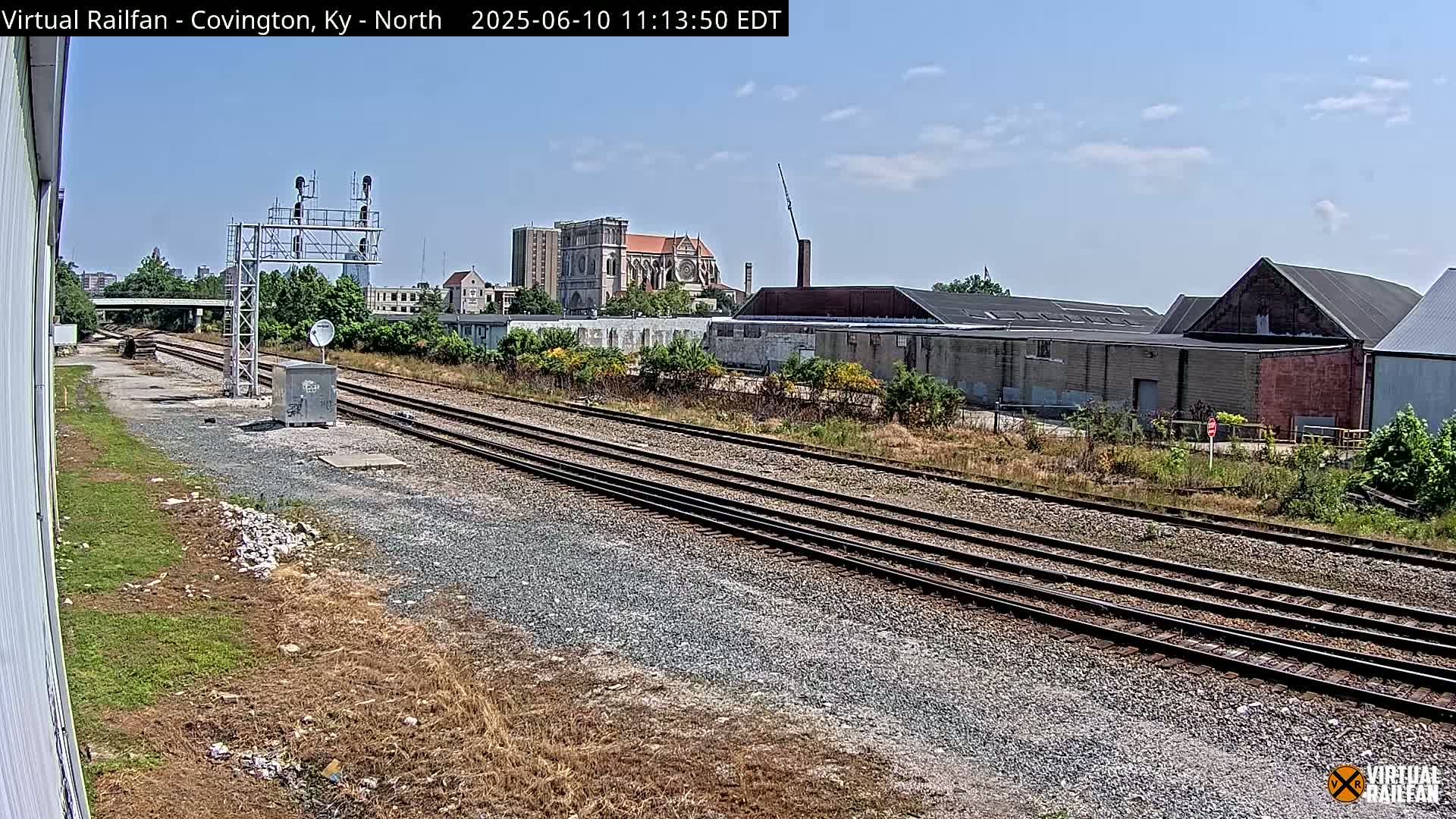 A view of multiple train tracks on a sunny day, with industrial buildings and a large church visible in the background.