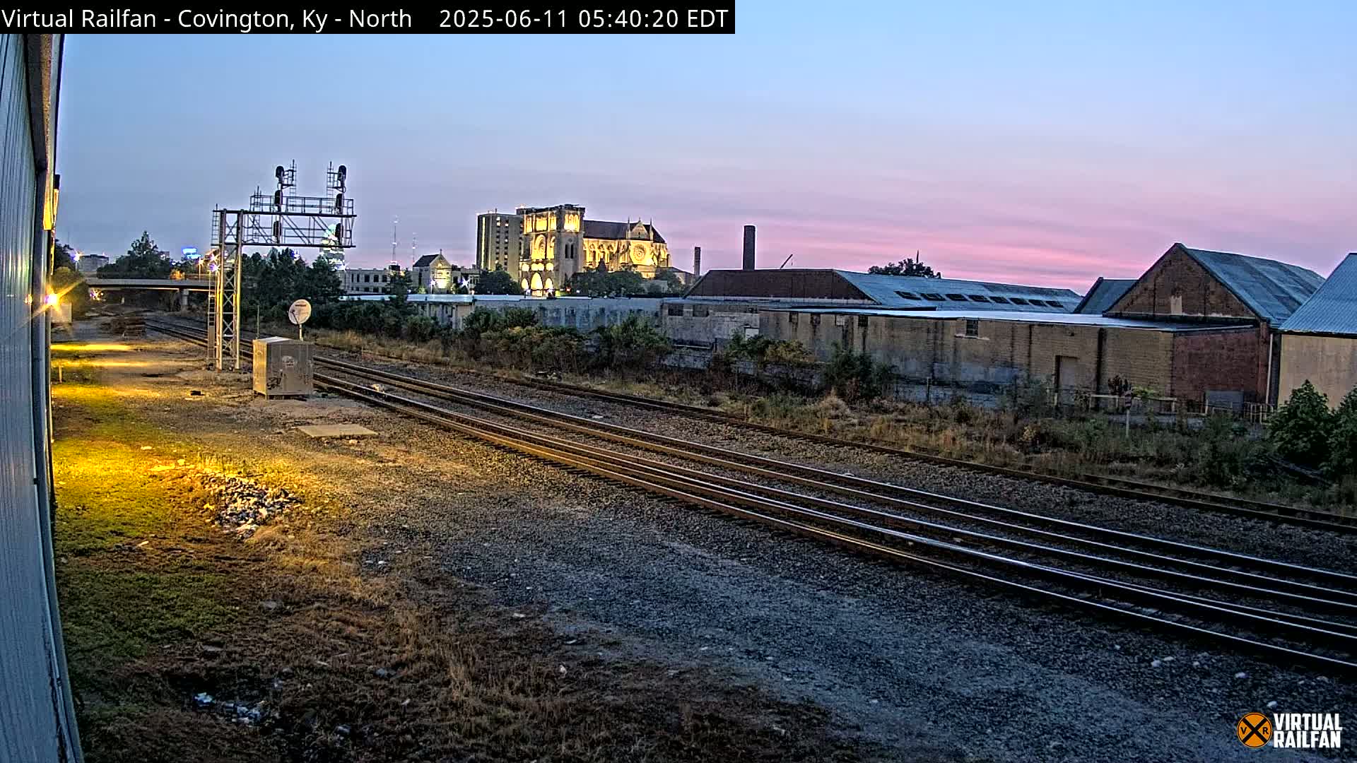 Multiple sets of train tracks stretch towards a city skyline at twilight, with a large cathedral visible in the distance under a pinkish-purple sky.