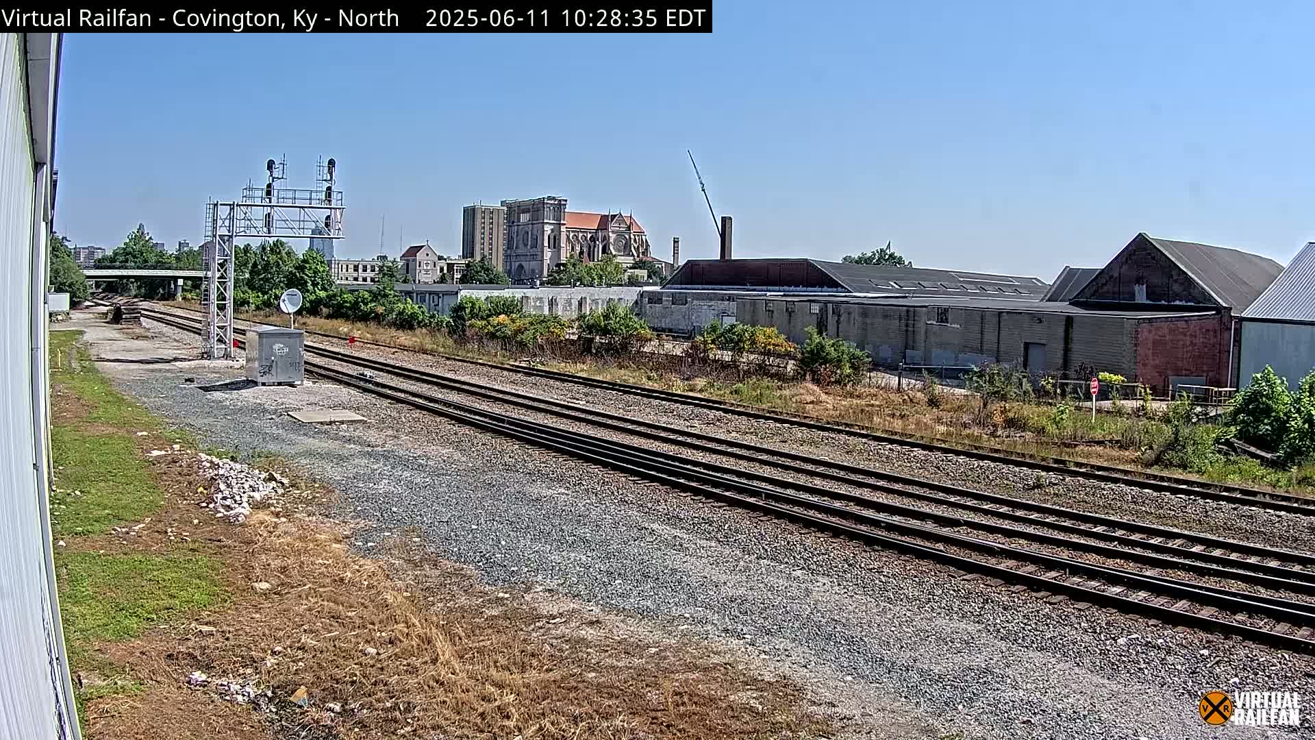 Multiple sets of train tracks extend into the distance under a clear blue sky, past industrial buildings and a large church.