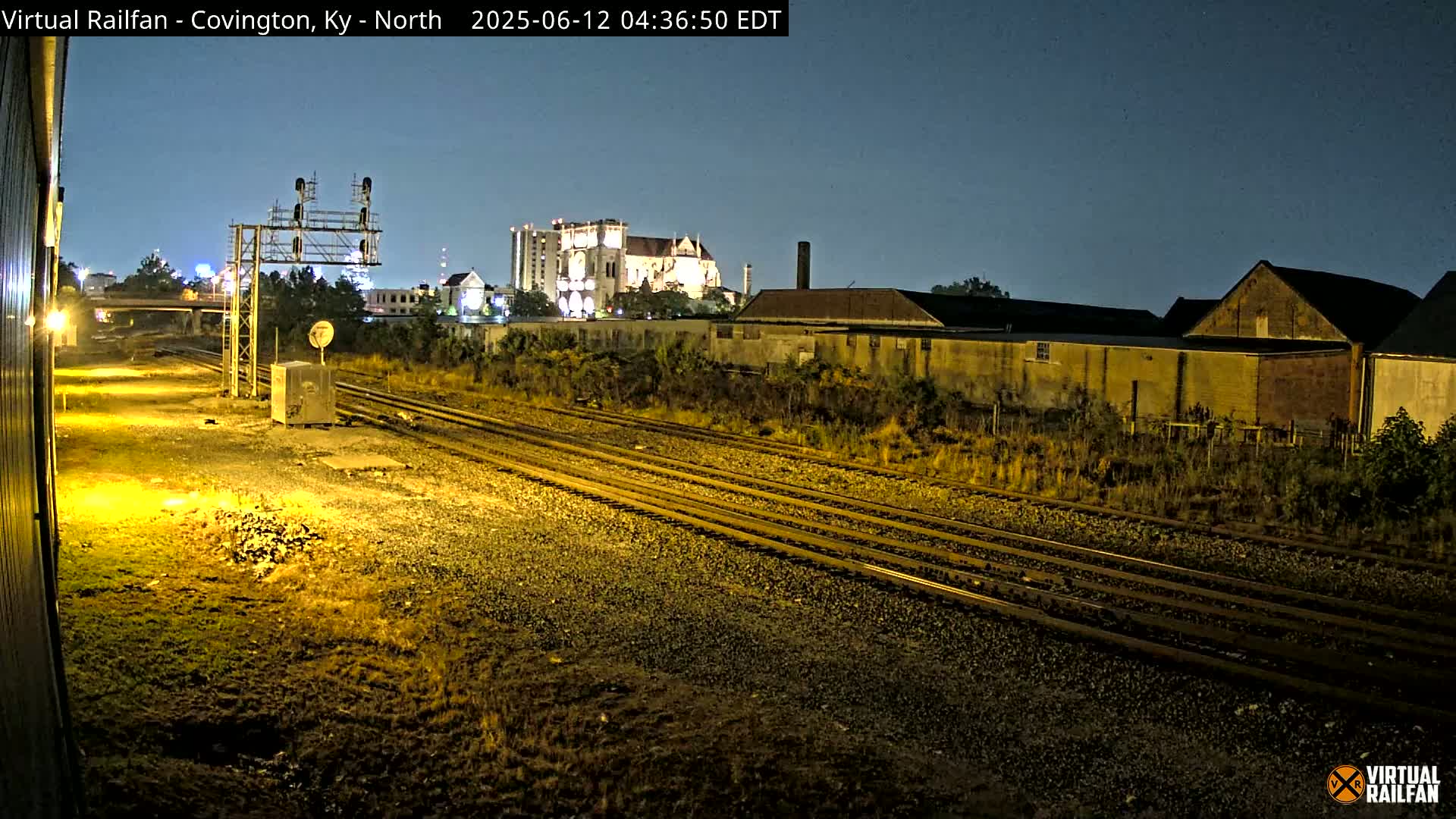 At night, under a clear sky, multiple train tracks extend toward a cityscape featuring buildings and a large structure that resembles a cathedral.
