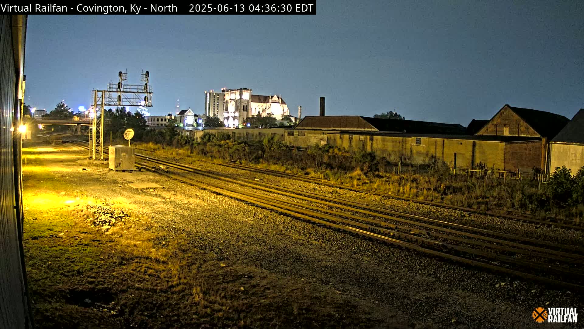 A nighttime view of multiple train tracks, industrial buildings, and a large building in the distance under a clear, dark sky.