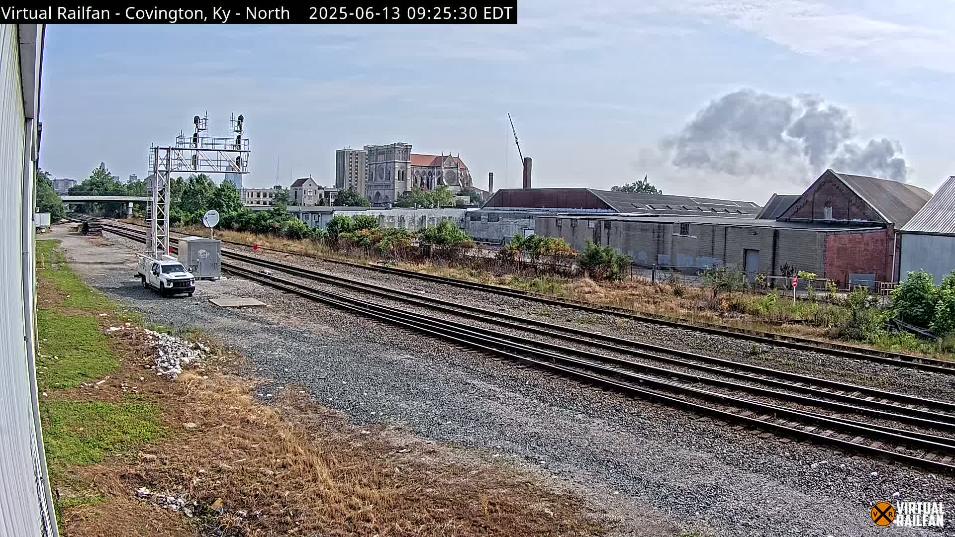 A view of multiple train tracks on a partly sunny day, with industrial buildings, a church, and a white truck visible in the background.