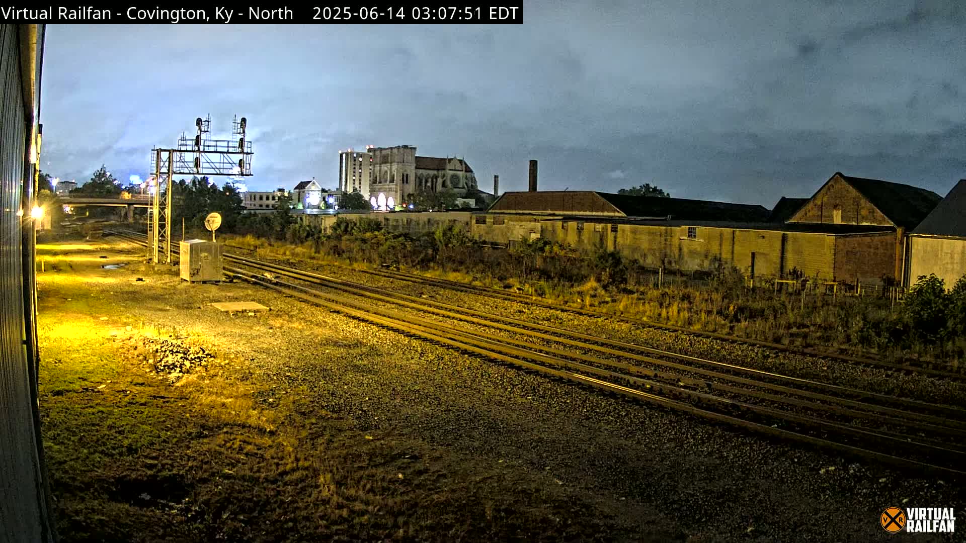 Multiple sets of train tracks at night under a cloudy sky, with buildings and vegetation in the background.