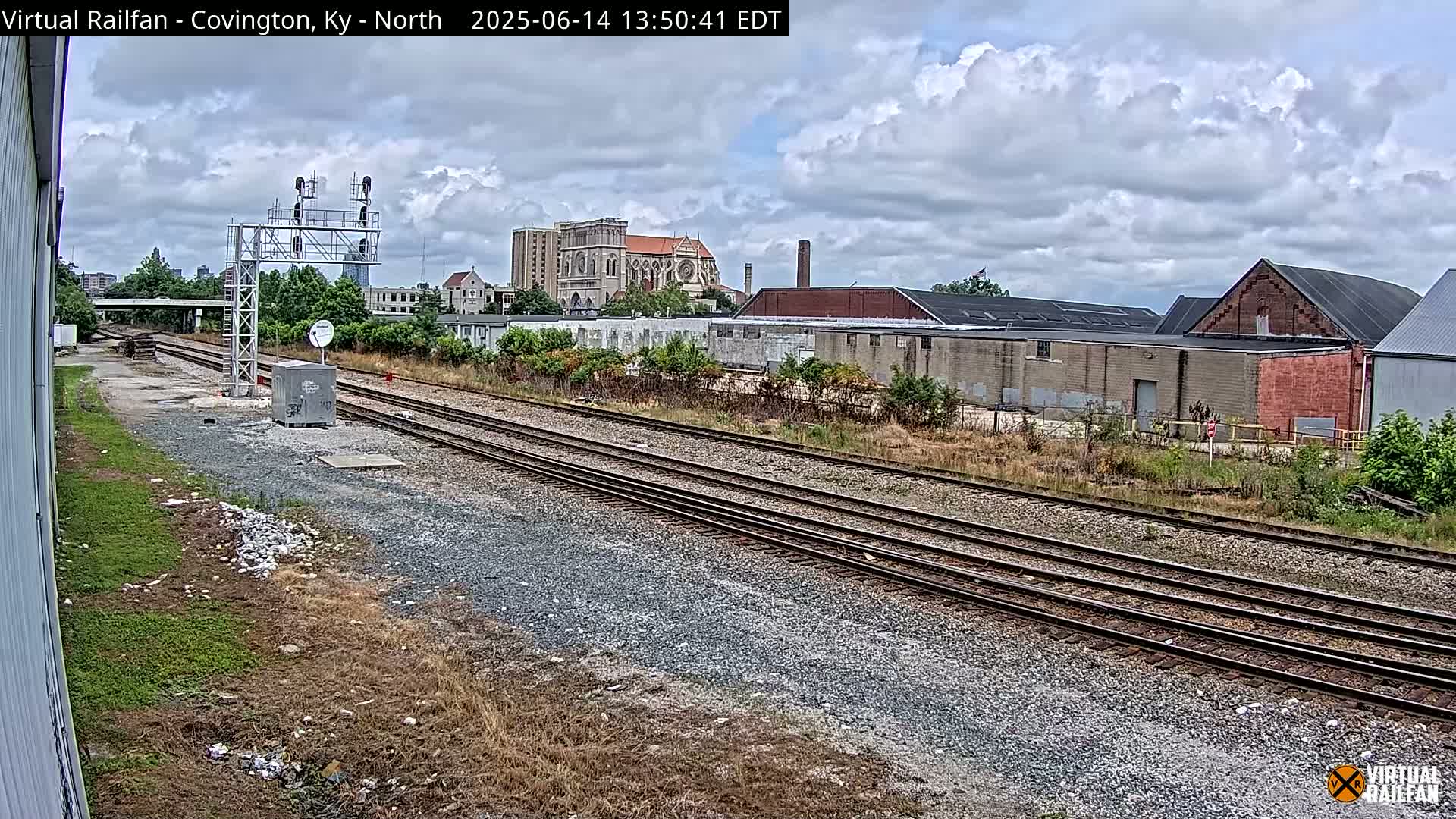 Multiple train tracks stretch into the distance under a partly cloudy sky, past industrial buildings and a large church.