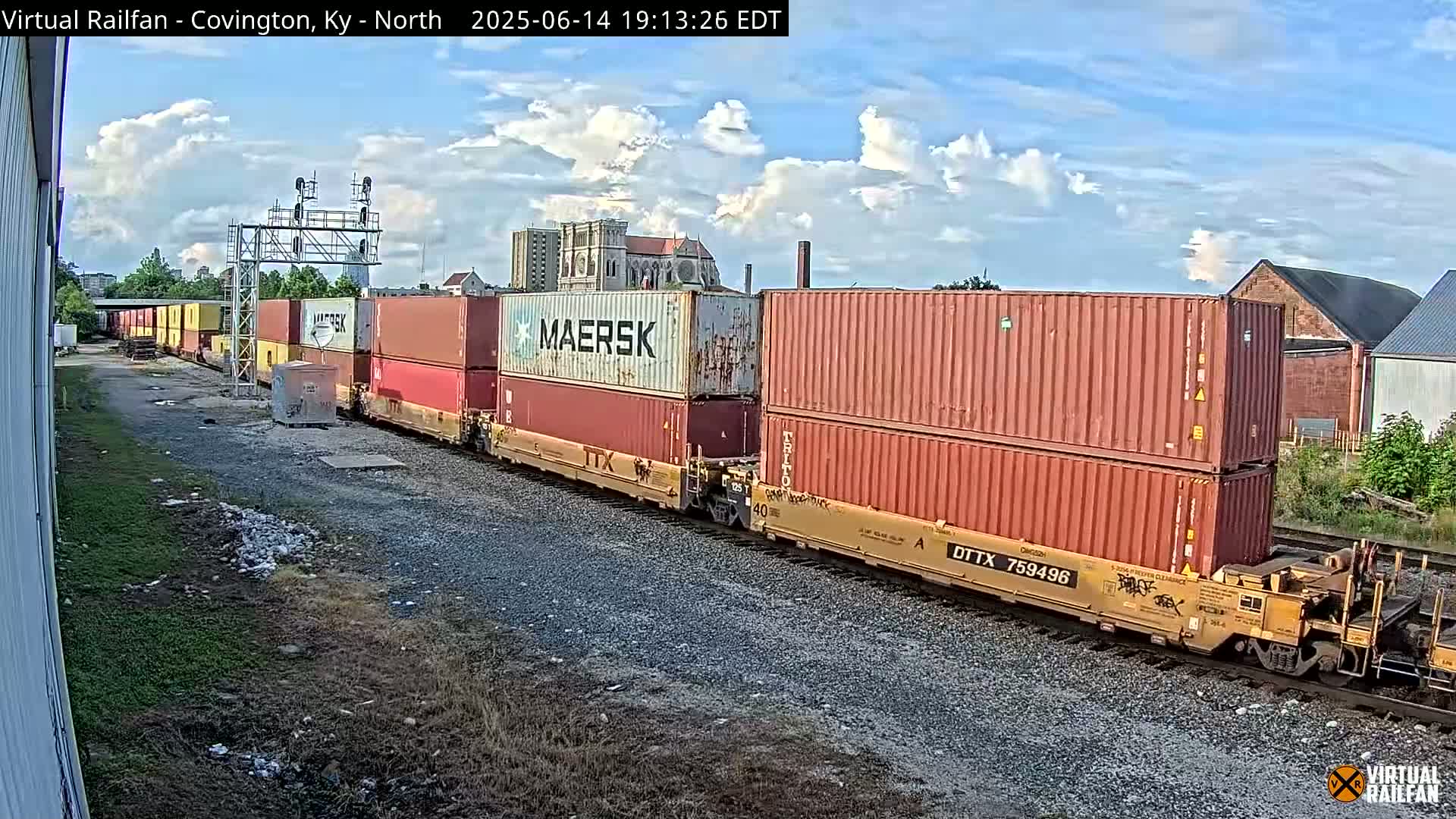 Multiple train tracks stretch into the distance under a partly cloudy sky, past industrial buildings and a large church.
