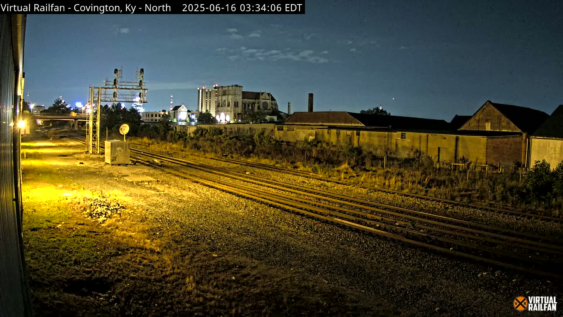 Under a dark, clear night sky, multiple train tracks curve through a mostly empty, grassy lot next to several buildings, including a large cathedral.