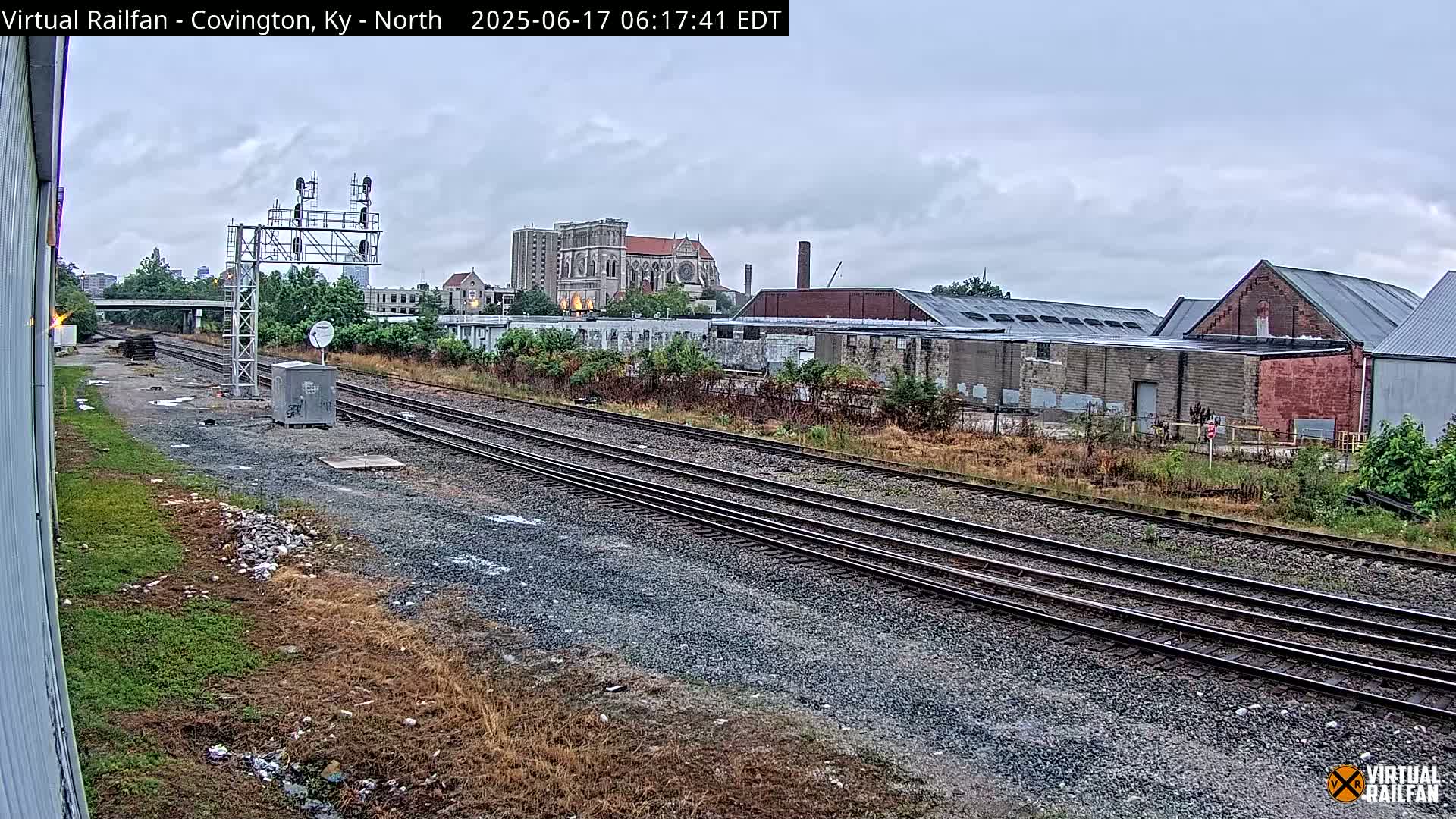 Multiple train tracks extend into the distance under an overcast sky, past industrial buildings and a large stone church.