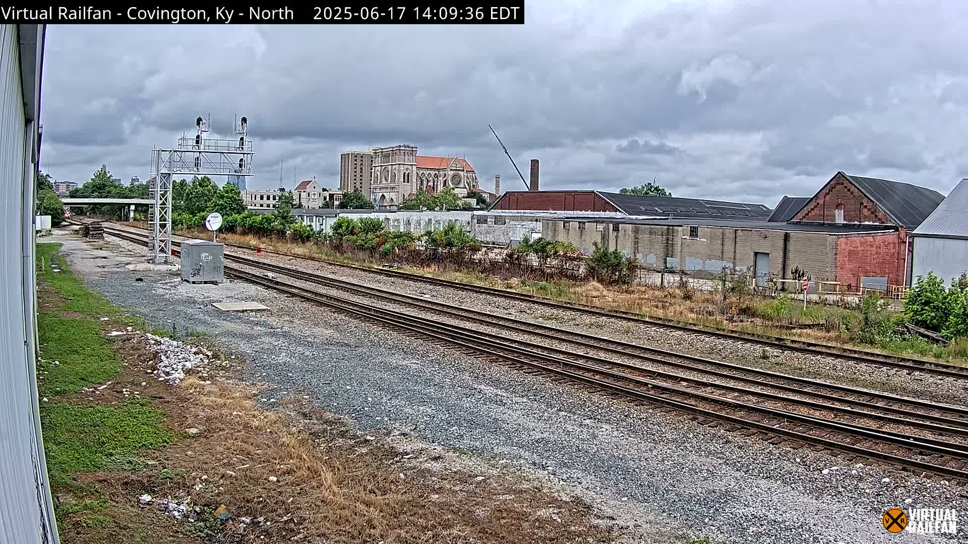 Multiple sets of train tracks stretch into the distance under an overcast sky, past industrial buildings and a large church.