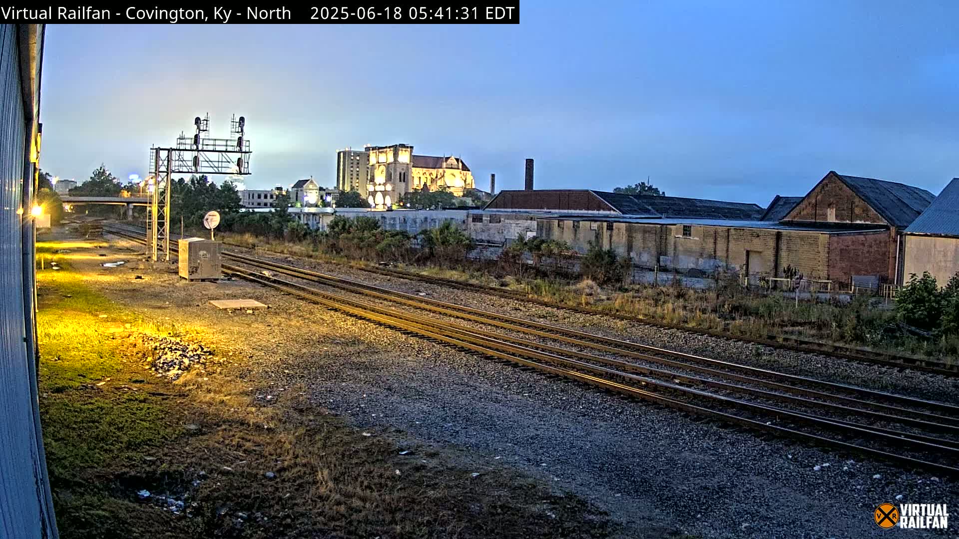 Under a twilight sky, three sets of train tracks stretch toward a cityscape featuring a large illuminated building, industrial structures, and overgrown vegetation alongside the tracks.
