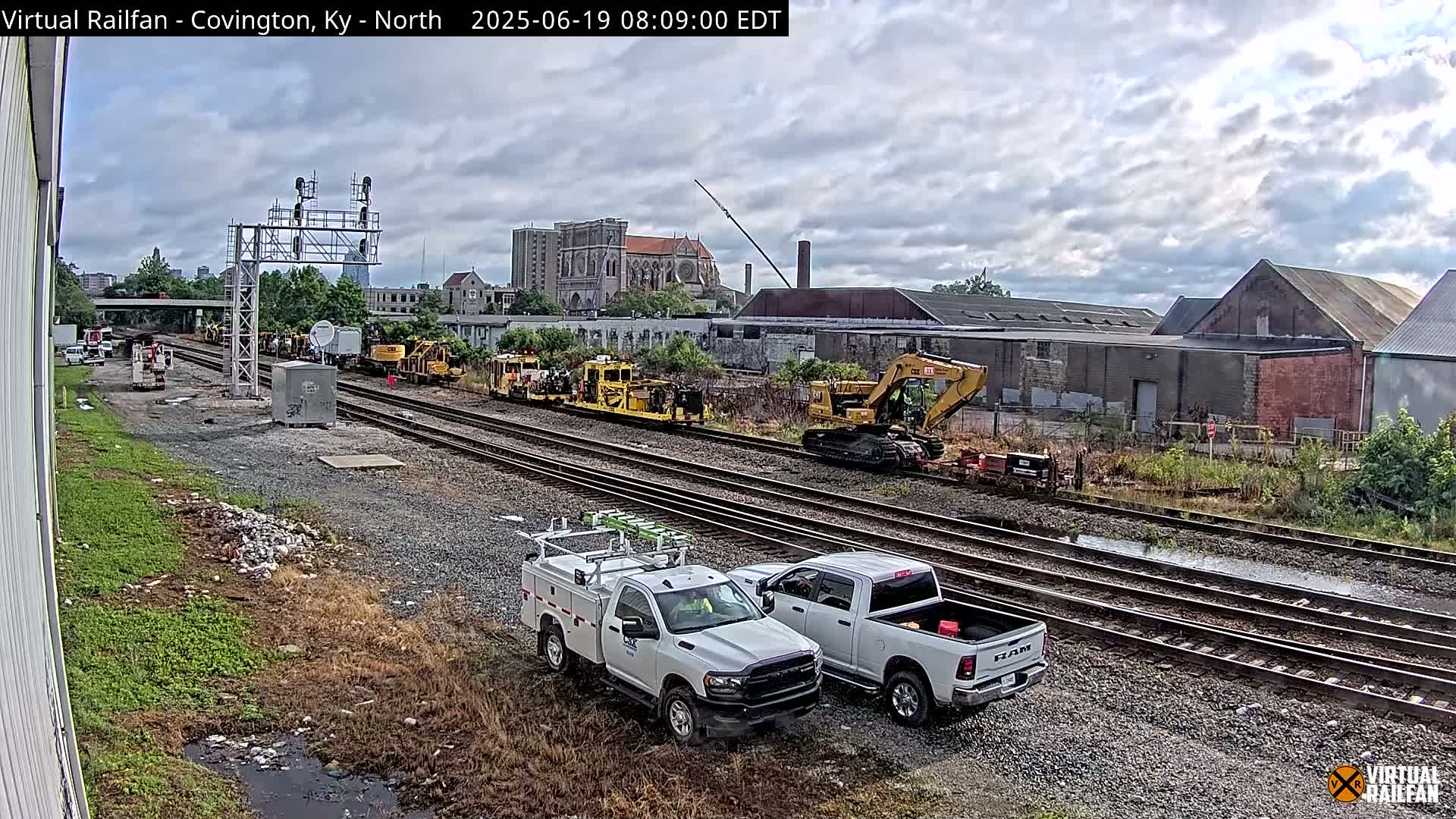 Under a cloudy sky, several pieces of yellow railway maintenance equipment are parked on a set of train tracks next to two white pickup trucks.