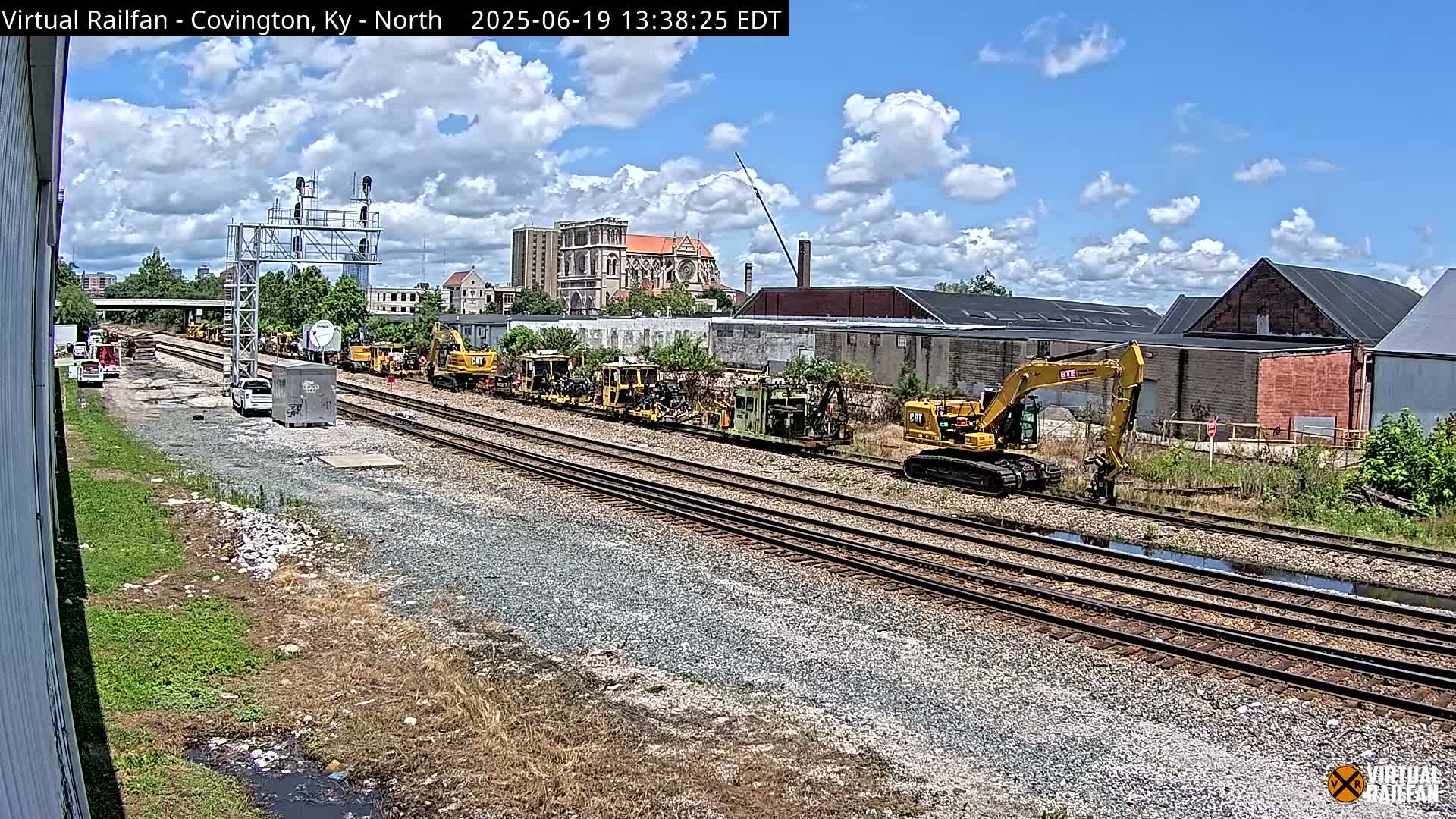 A partly cloudy day reveals multiple yellow construction vehicles parked along railroad tracks near industrial buildings and a church in the background.