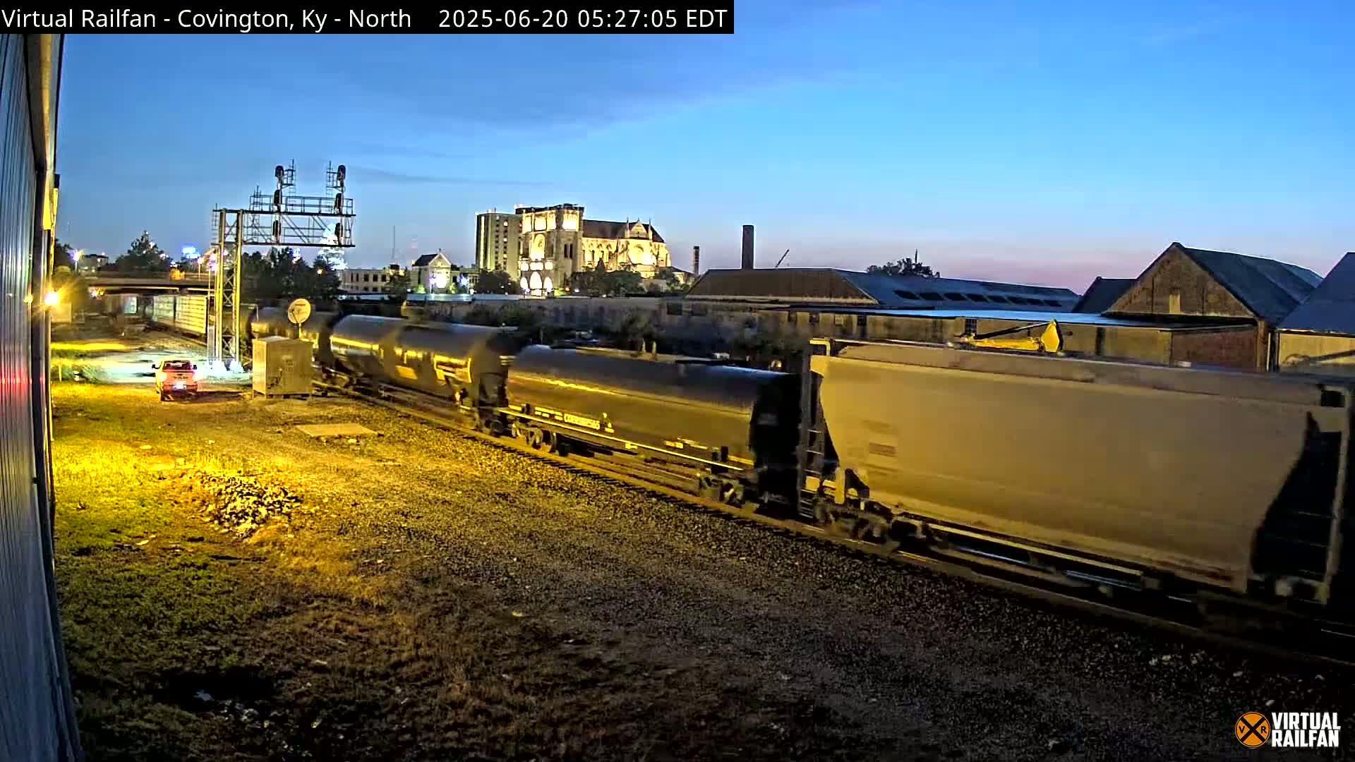 A long freight train passes by under a twilight sky, with a vehicle visible in the foreground.