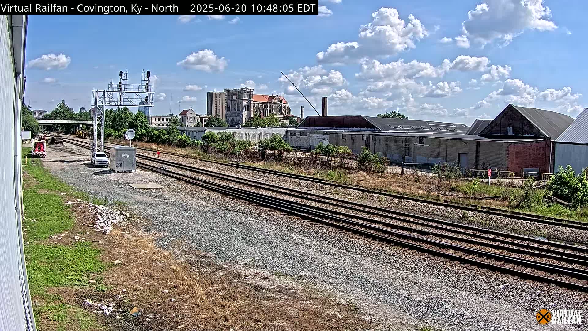 A sunny day with partly cloudy skies shows multiple train tracks leading toward buildings and a large cathedral in the distance.