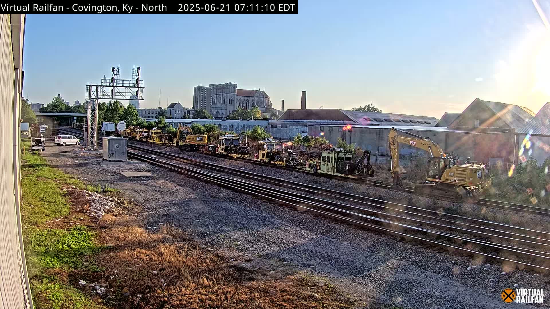 Under a clear, sunny sky, several pieces of yellow railway maintenance equipment are parked along multiple train tracks next to a building.