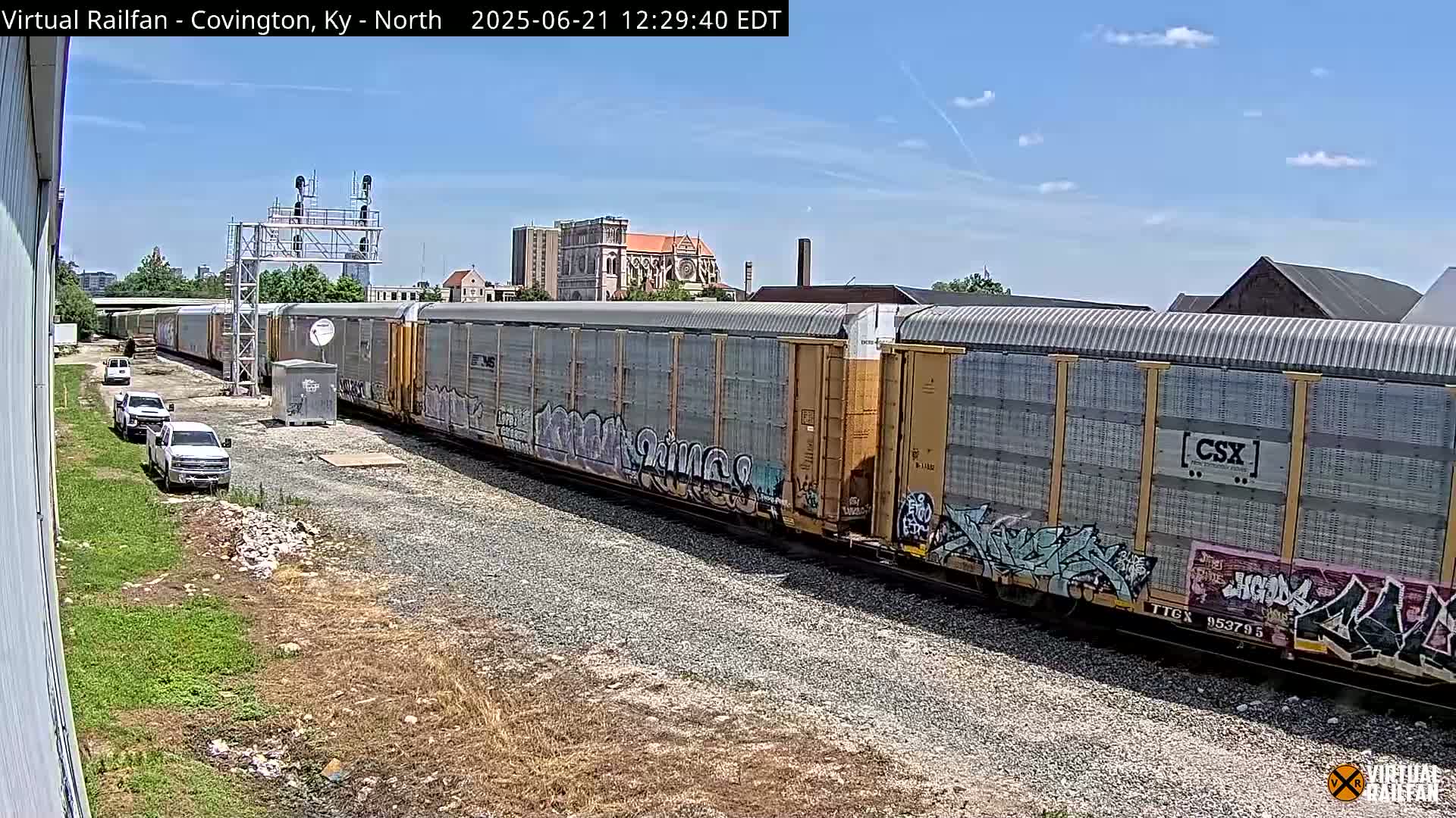 A long freight train with graffiti on some cars sits on tracks under a mostly sunny sky, with several white pickup trucks parked nearby.