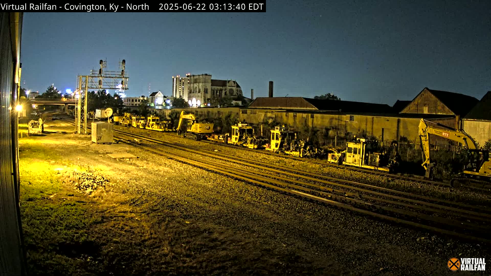 Under a dark night sky, several pieces of yellow railway maintenance equipment are parked along multiple sets of train tracks near buildings.