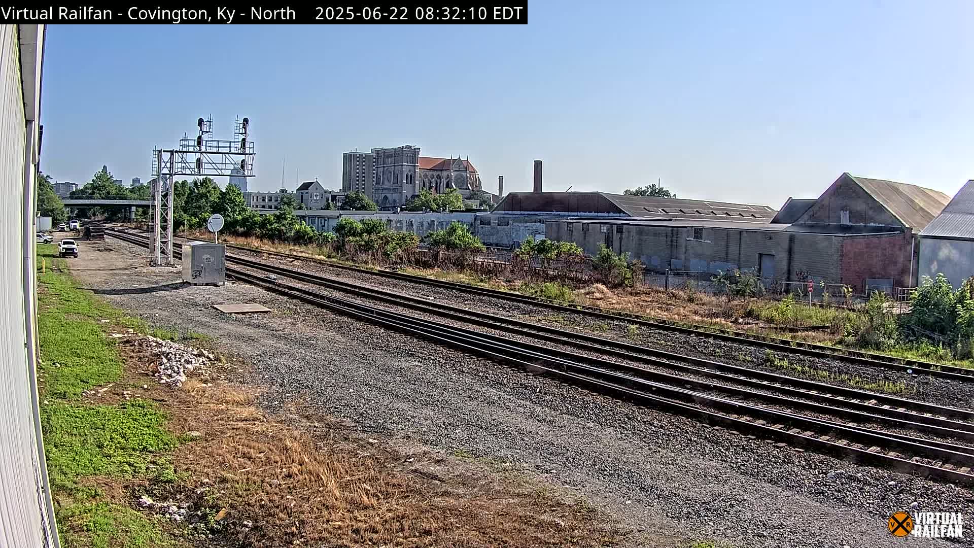 A view of multiple train tracks on a sunny day, with industrial buildings and a church visible in the background.