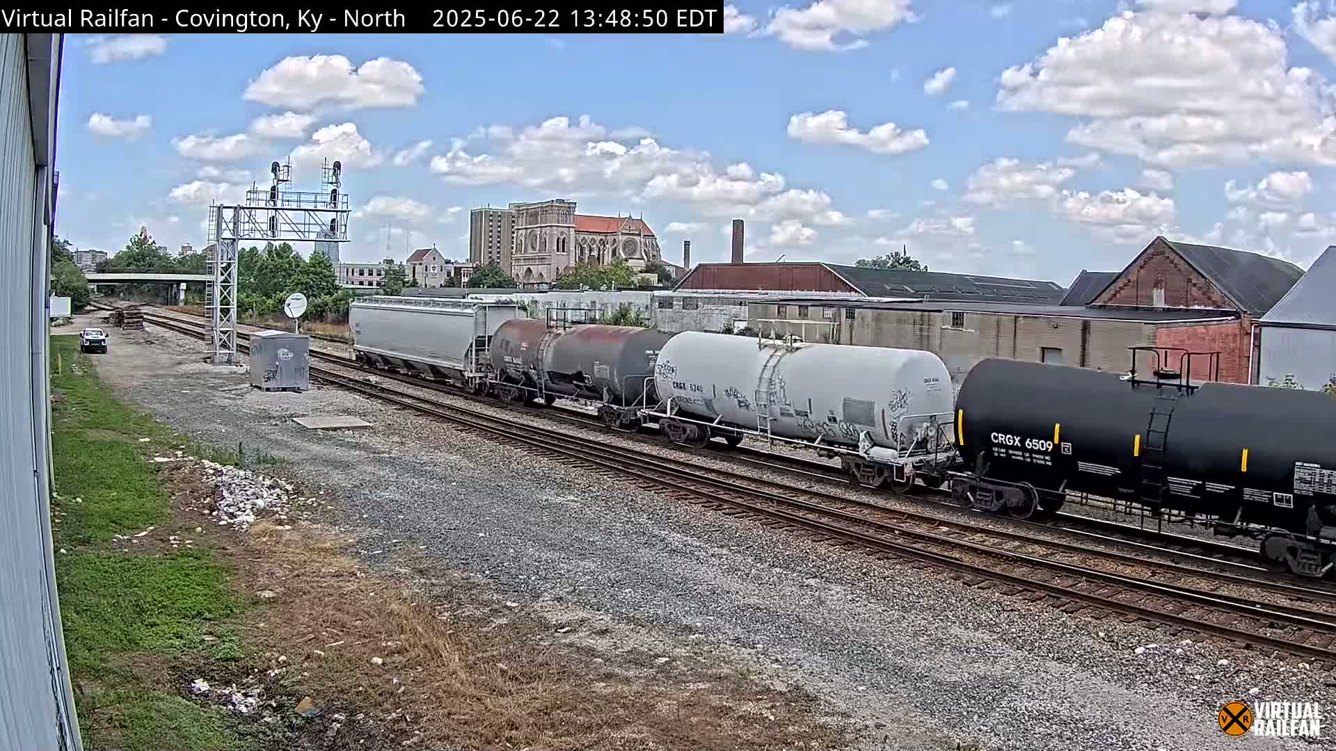 A freight train carrying several tanker cars moves along a set of railroad tracks under a partly cloudy sky.