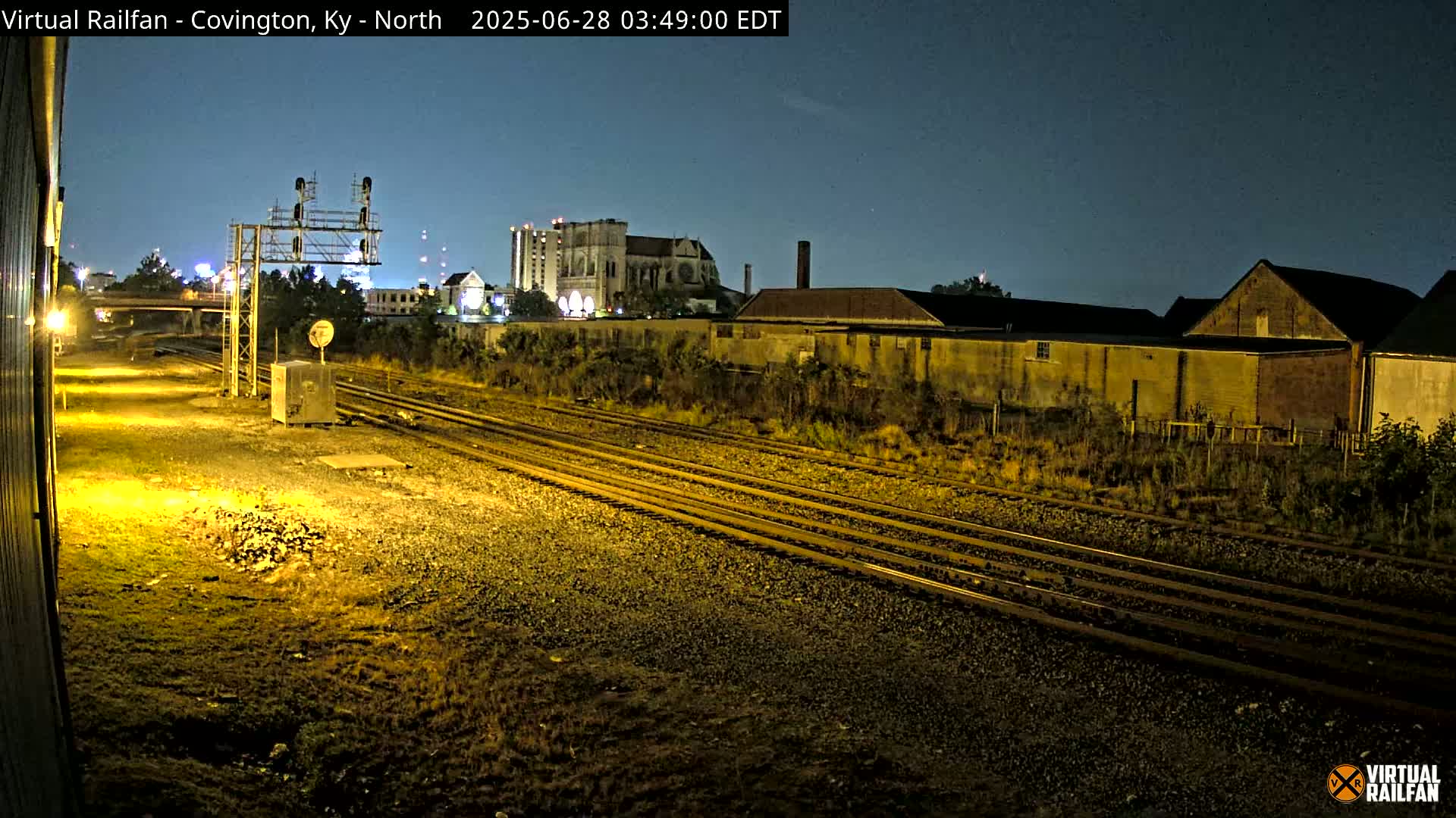 Under a clear night sky, multiple train tracks extend into the distance past industrial buildings and a large cathedral.