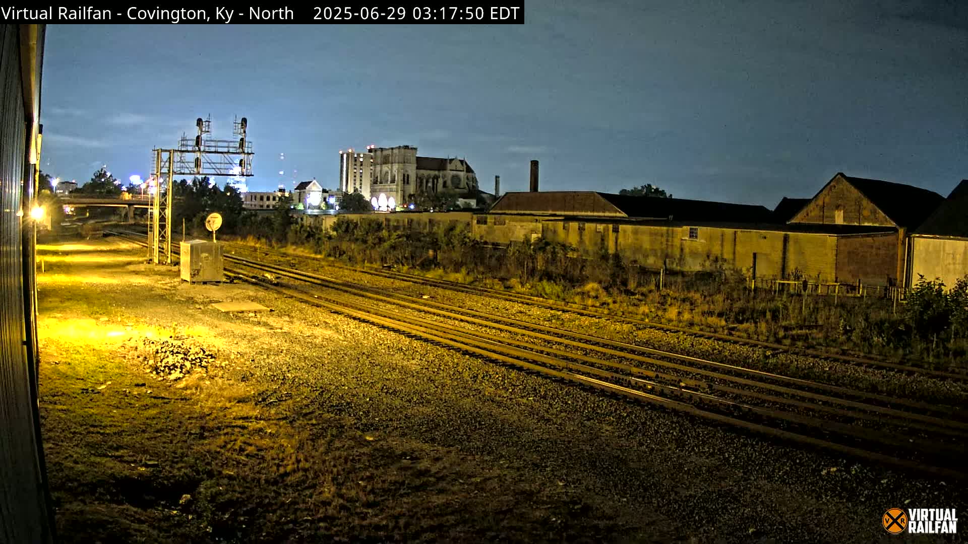 At night, under a twilight sky, multiple sets of train tracks extend into the distance past industrial buildings and a large church.