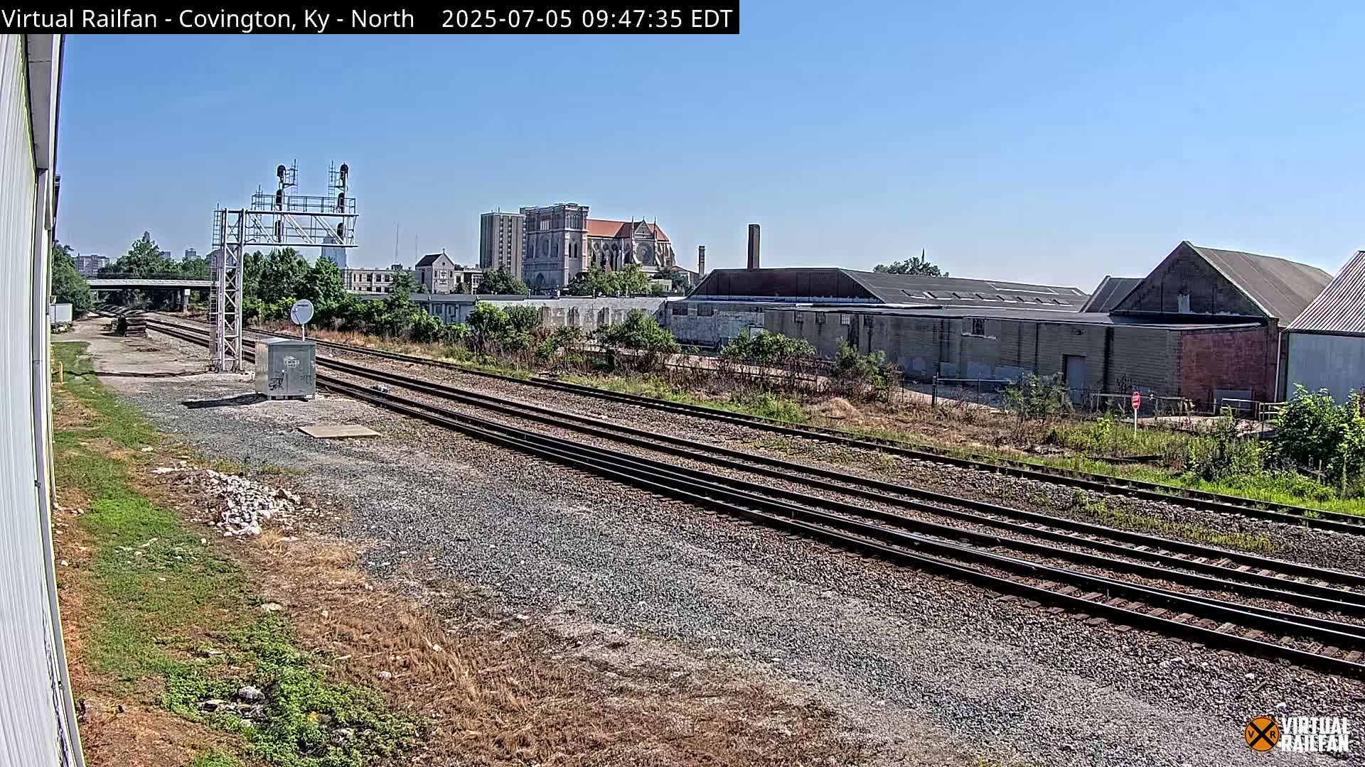 Multiple sets of train tracks extend into the distance under a clear blue sky, past industrial buildings and a church.