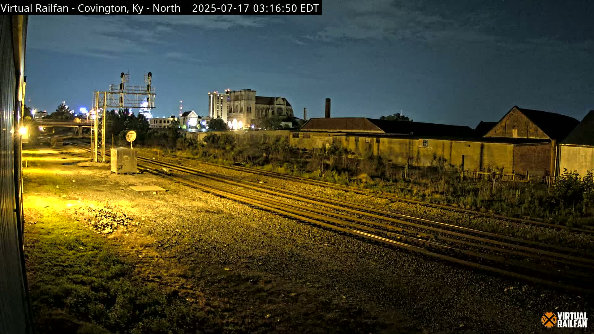At night, multiple sets of train tracks extend into the distance past some buildings under a mostly clear sky.