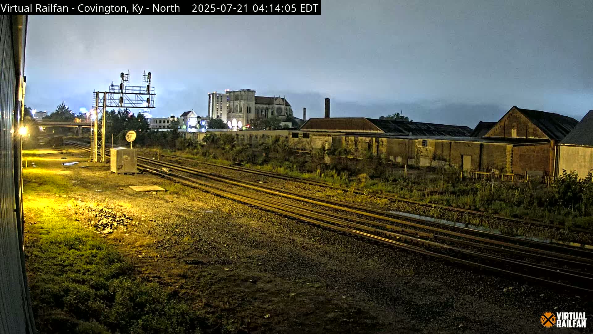 A nighttime view of several sets of train tracks running past industrial buildings and a cathedral under a cloudy sky.