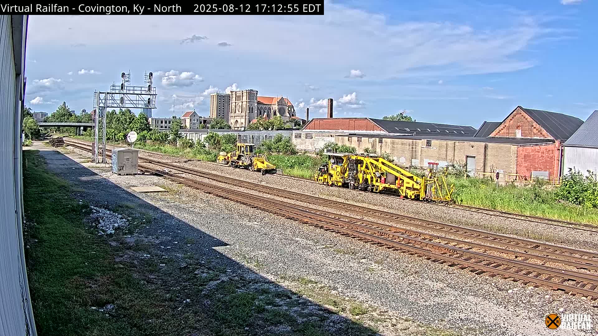 Two yellow railway maintenance vehicles are parked on a set of train tracks on a sunny day, with industrial buildings and a large cathedral visible in the background.