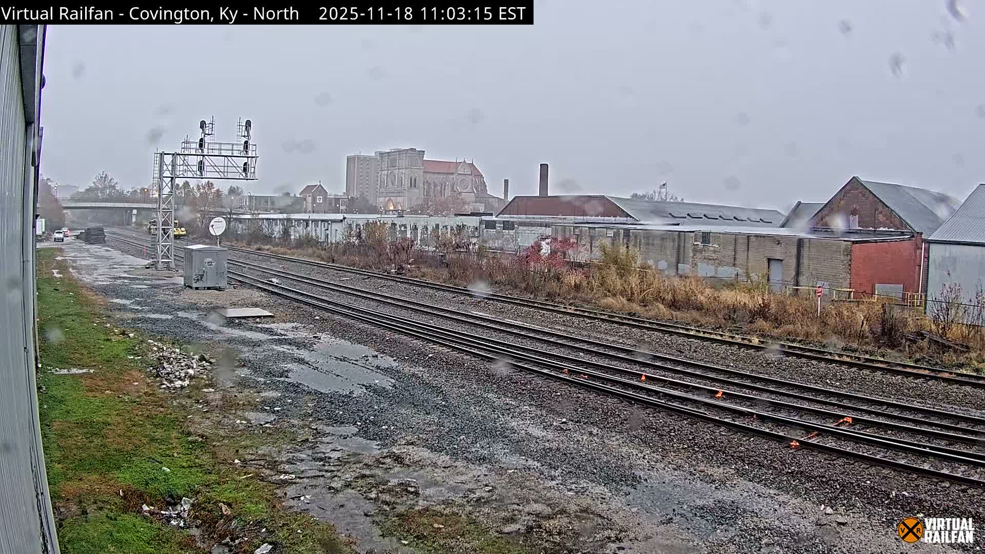 Two yellow railway maintenance vehicles are parked on a set of train tracks on a sunny day, with industrial buildings and a large cathedral visible in the background.