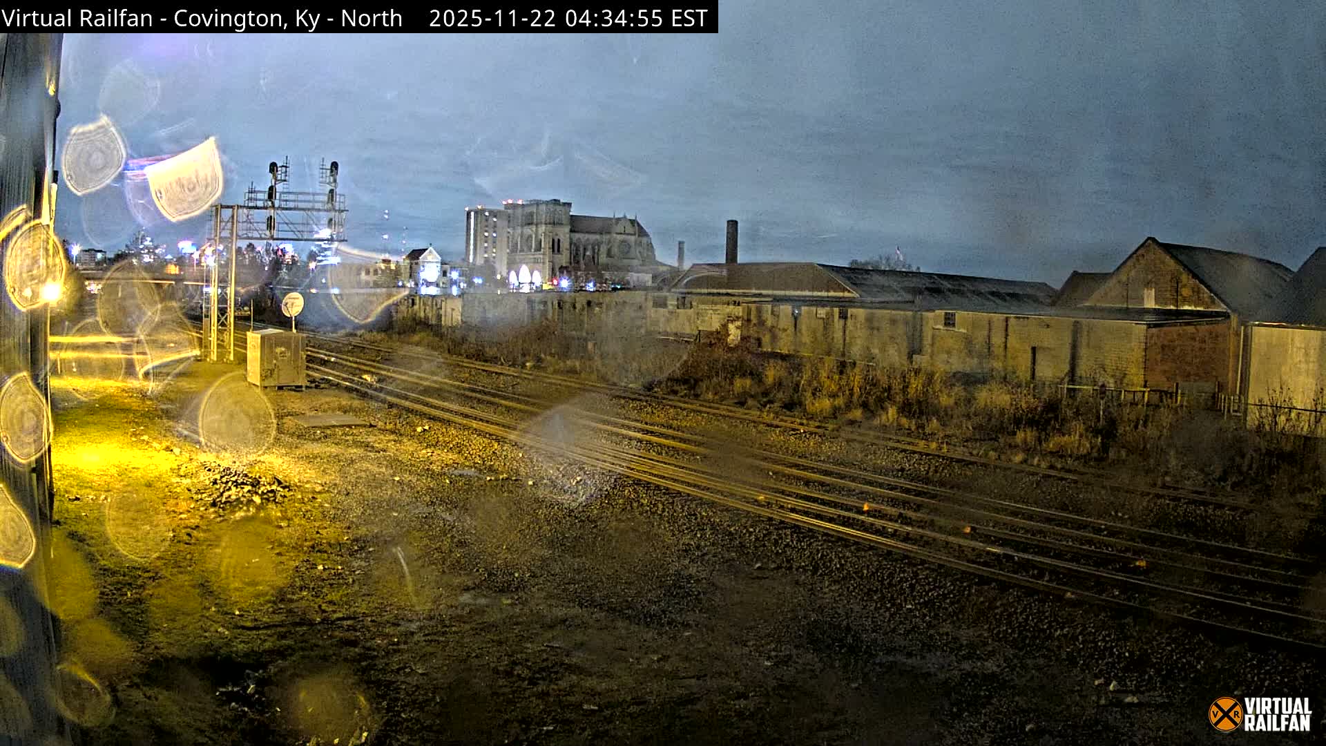 A rain-streaked camera lens captures an urban rail yard at night, with multiple tracks leading past industrial buildings towards a prominent ornate building and city lights in the distance under a dark sky.