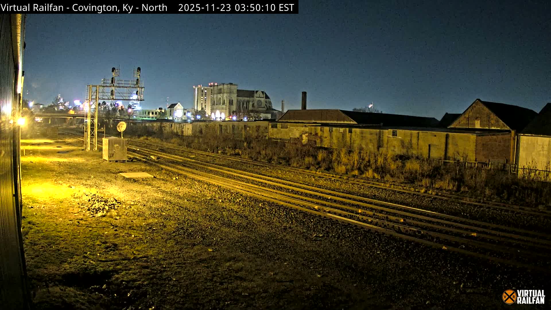 An illuminated railway yard with multiple tracks and signals is visible at night under a clear sky, with various buildings and a prominent cathedral-like structure in the background.