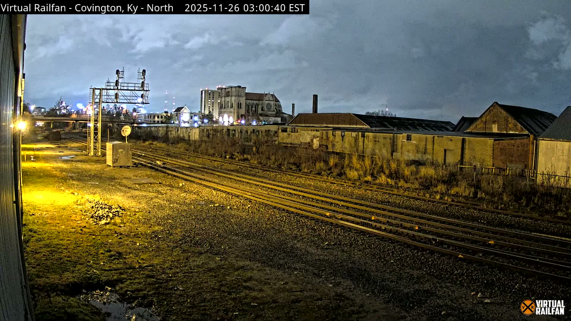 A nighttime view of an illuminated train yard reveals multiple tracks, industrial buildings, and a distant city skyline, all under a dark, overcast sky with puddles reflecting the dim light.