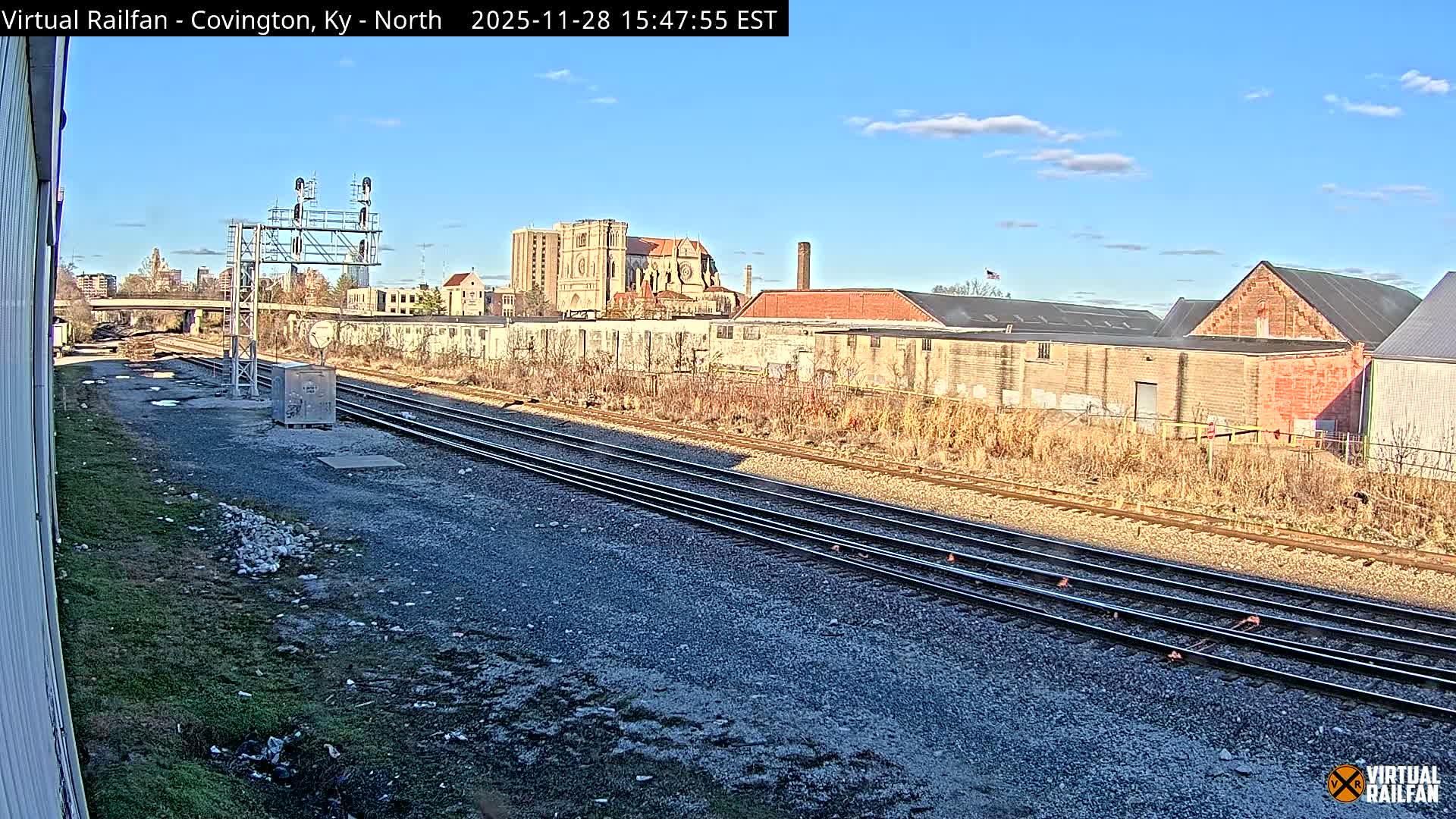 Multiple train tracks run through an urban setting under a clear and sunny sky with scattered clouds, flanked by industrial buildings and a distant ornate structure, with a signal gantry prominent in the midground.