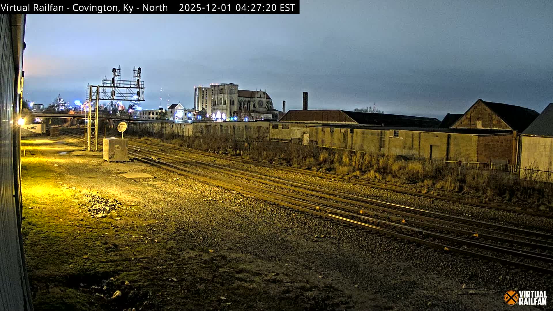 A nighttime scene reveals multiple railroad tracks stretching through an urban environment under an overcast sky, bordered by industrial buildings and a distant city skyline illuminated by artificial lights.